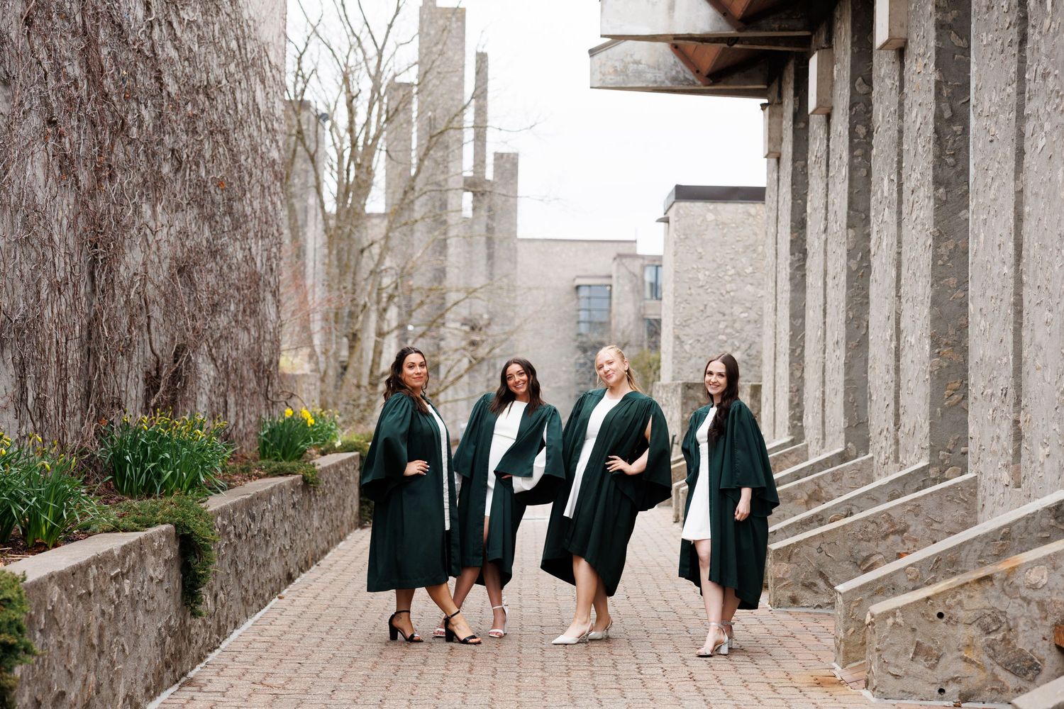 A group of graduates wearing dark green robes walk together along a stone pathway on a college campus.