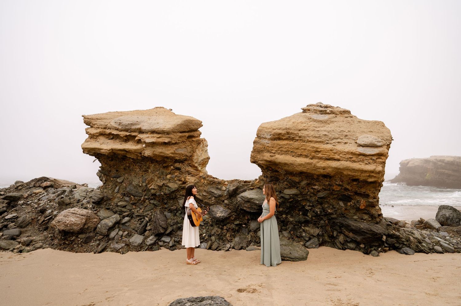 Two people stand between large rock formations on a misty beach.