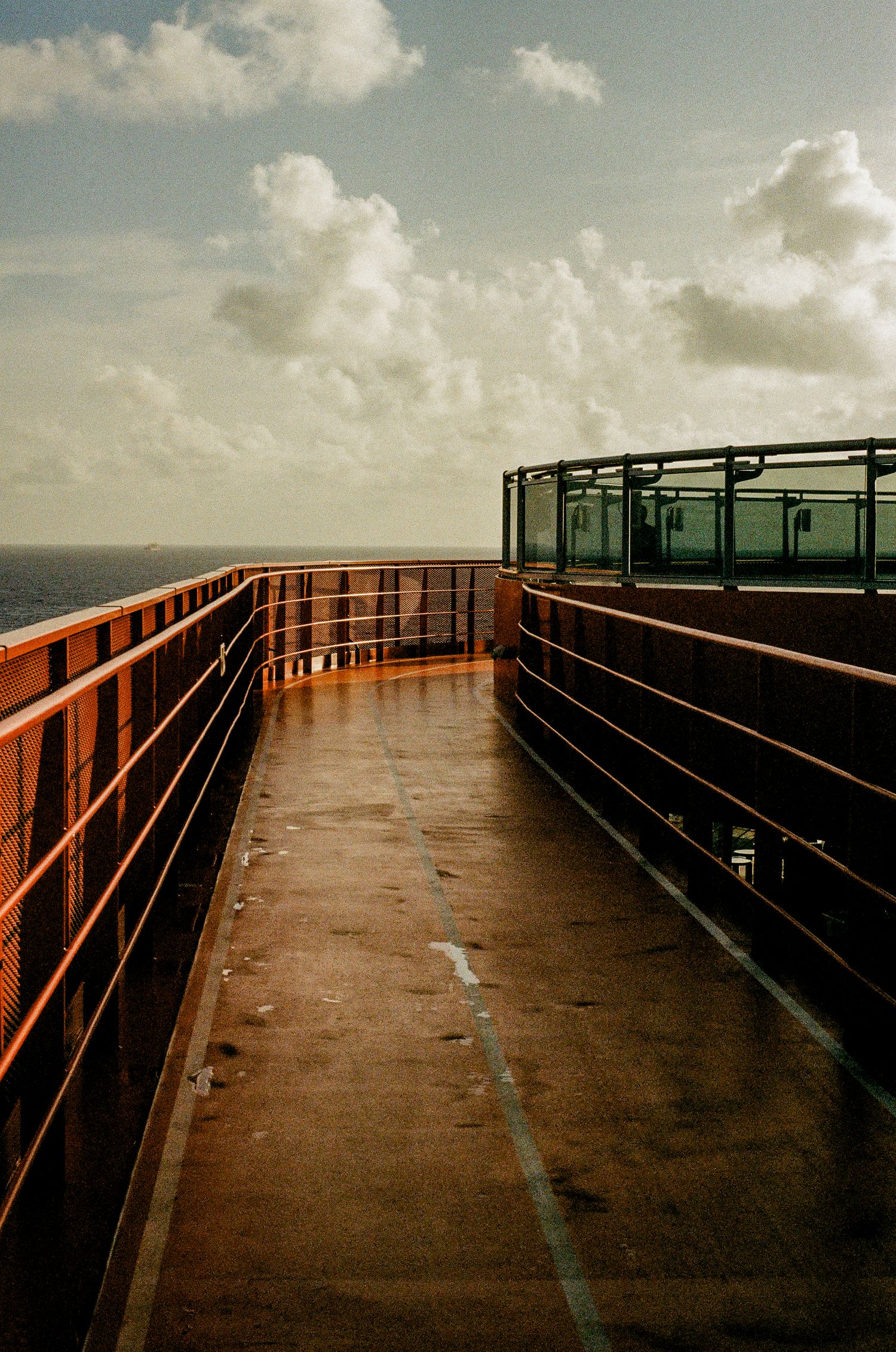 A curved walkway with rusted orange railings leading to a glass enclosure against a cloudy sky over the ocean.