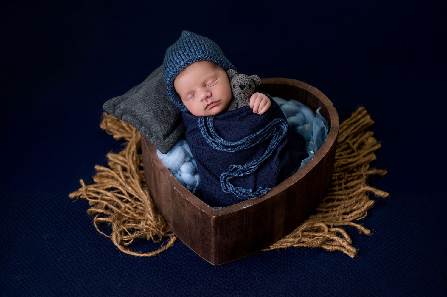 Sleeping infant wrapped in navy blue swaddle rests peacefully in wooden heart basket with rustic fringe trim against dark backdrop.