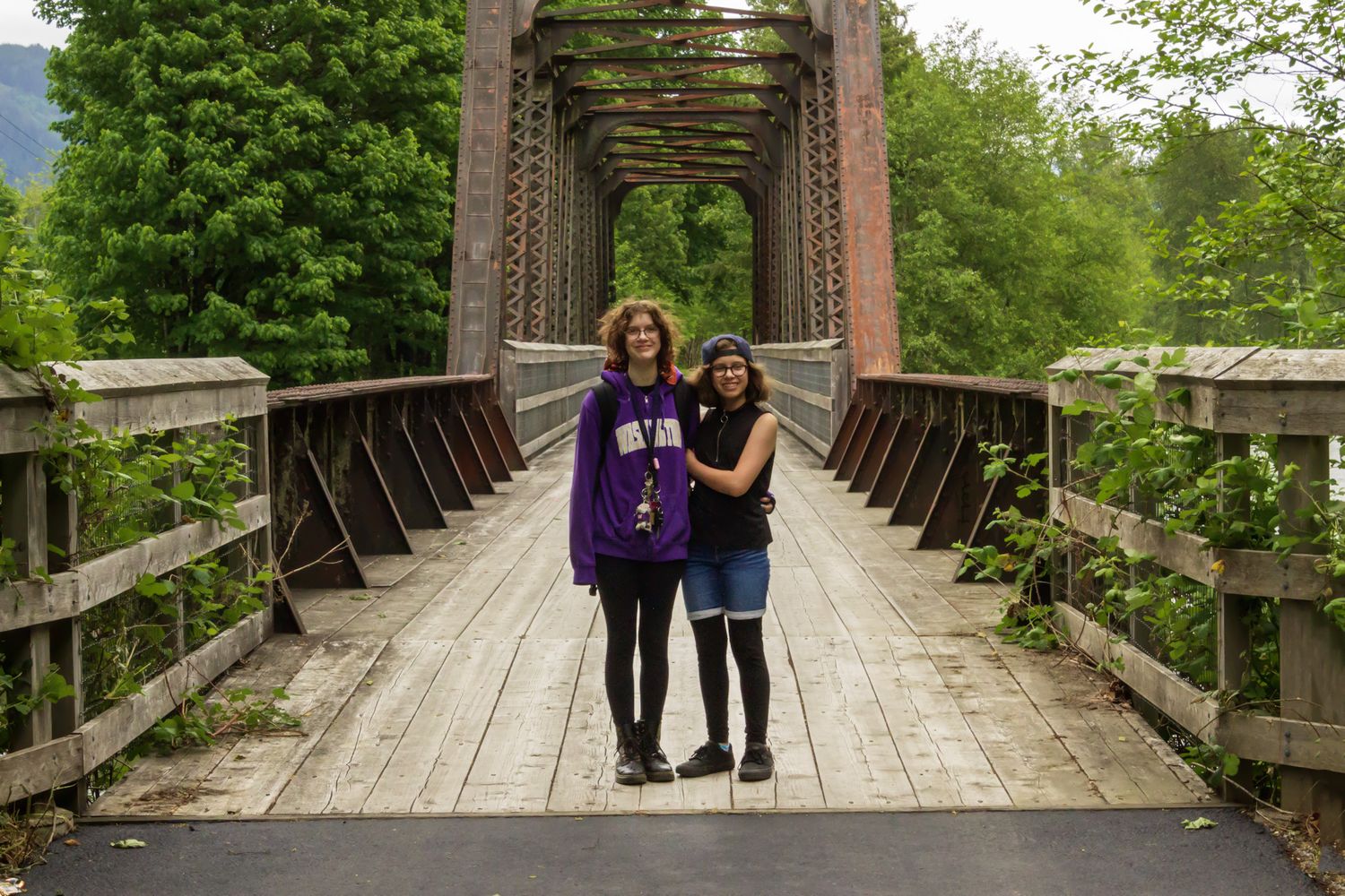 Two people standing together on a wooden railroad bridge surrounded by green trees and mountains.