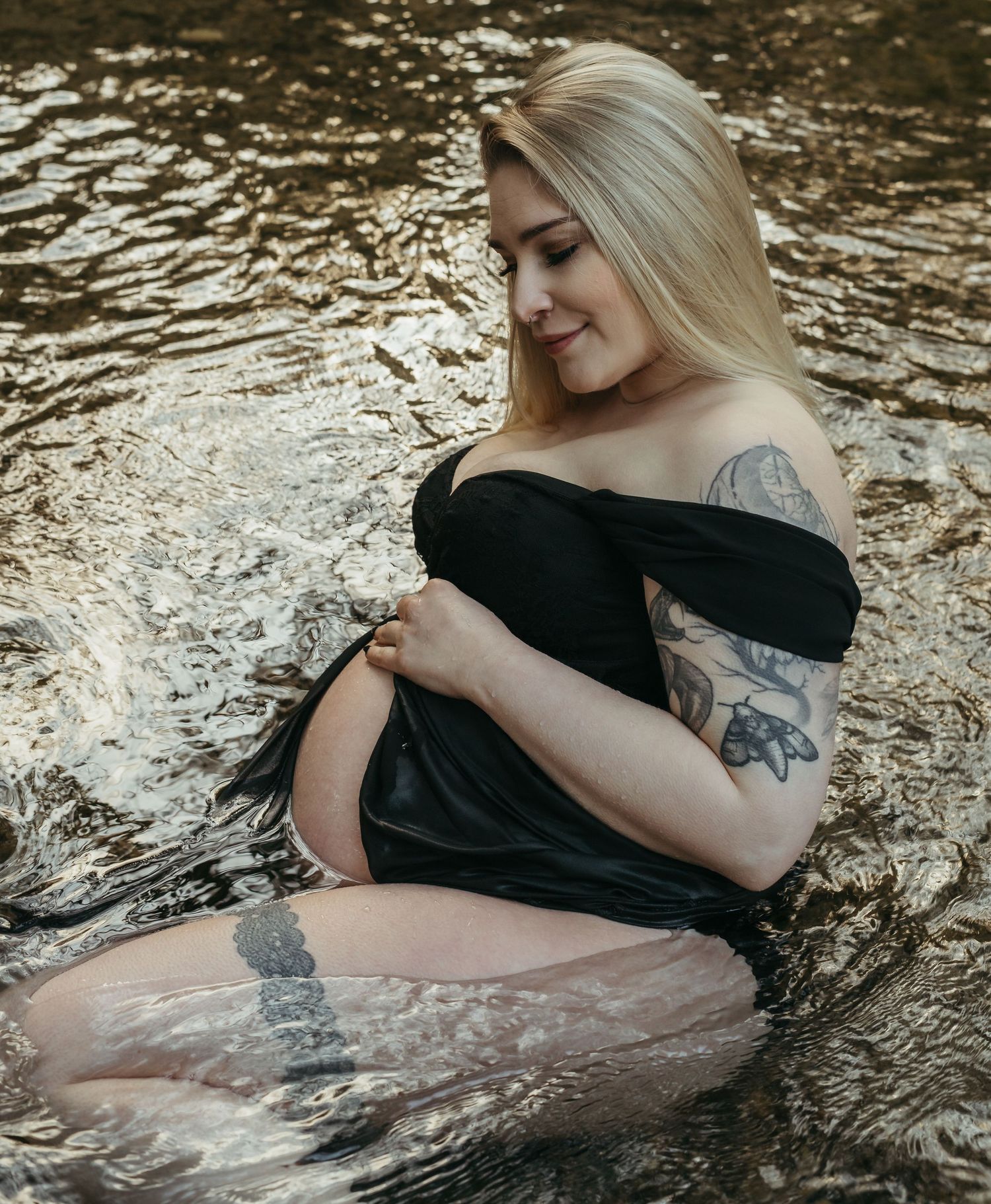 A maternity photo session in flowing water with a black off-shoulder dress and visible tattoos creates an ethereal mood.