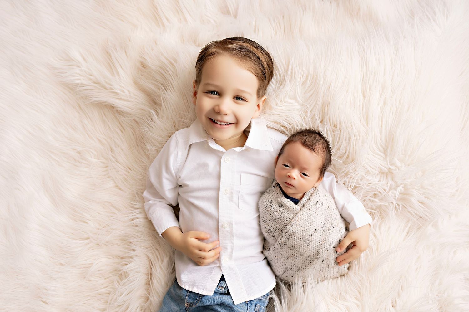 Two small children lying on a soft cream-colored fur rug, wearing light-colored clothing against a pale background.