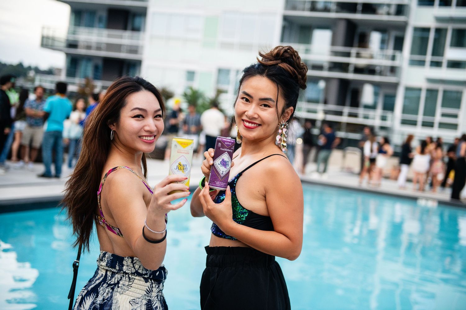 Two friends enjoying poolside drinks at rooftop party with city buildings in background.