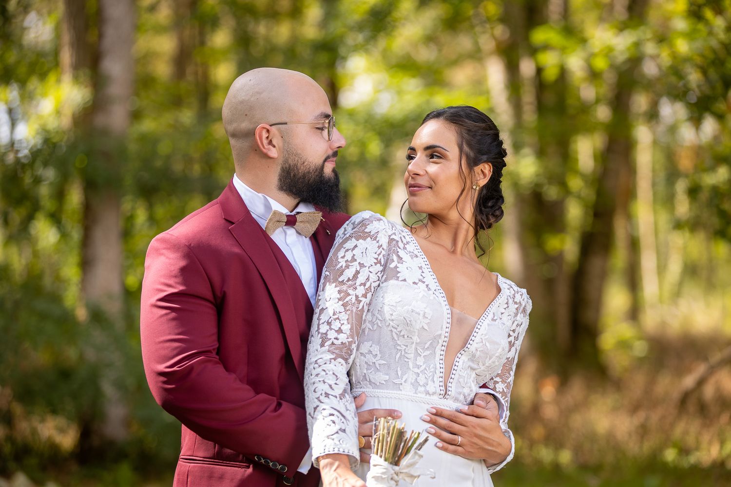 A romantic wedding portrait outdoors with sunlit forest backdrop and elegant attire.