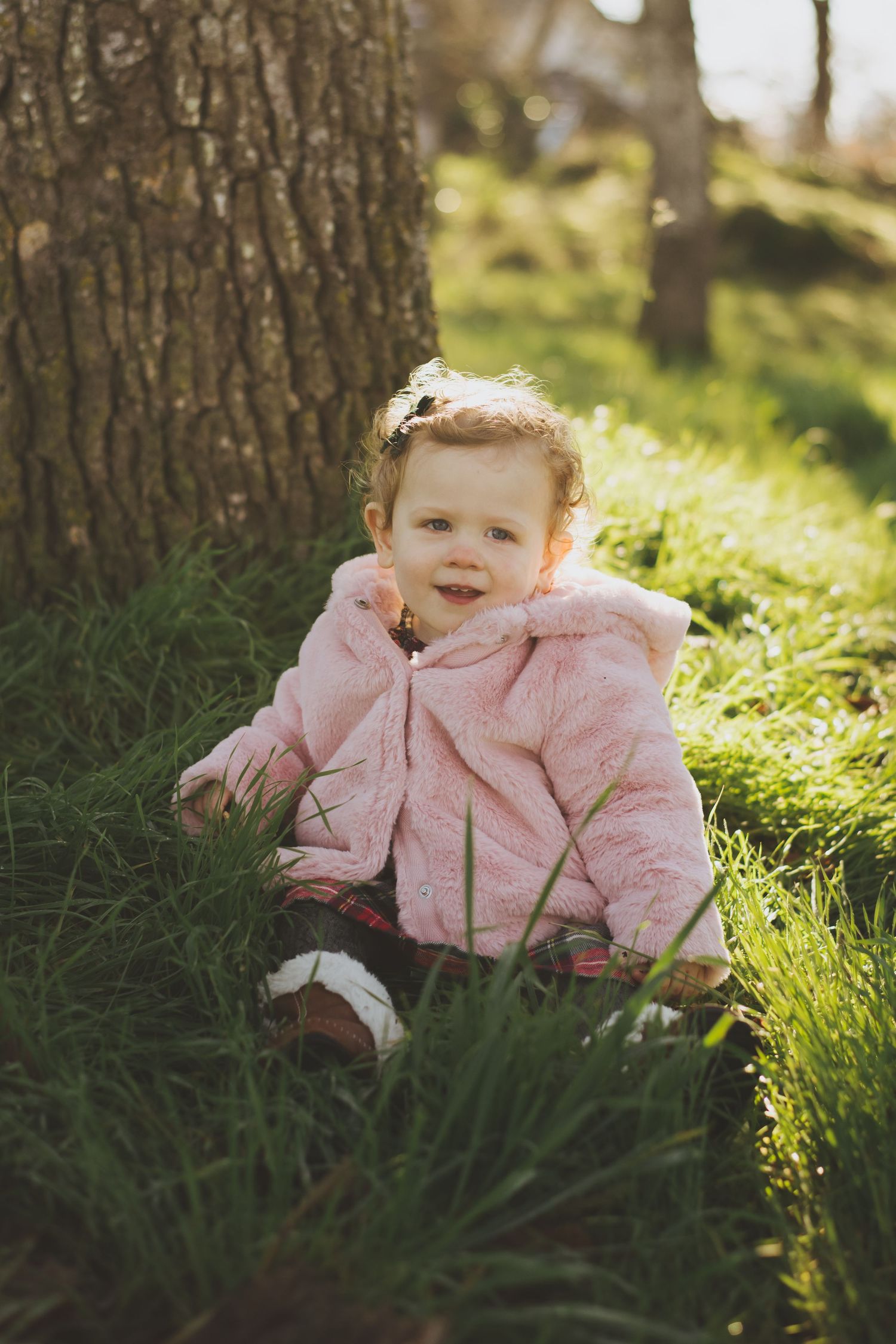 Baby girl wearing a pink fuzzy coat sits in green grass beside a tree trunk on a sunny day.