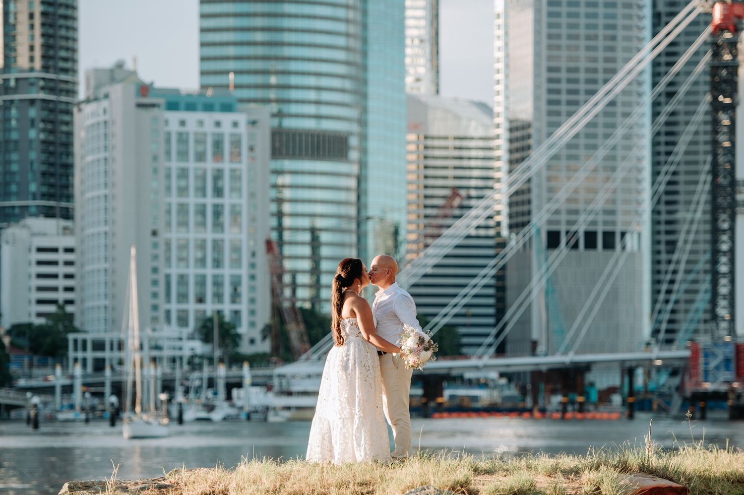 A newlywed couple poses against a modern cityscape backdrop with tall buildings.