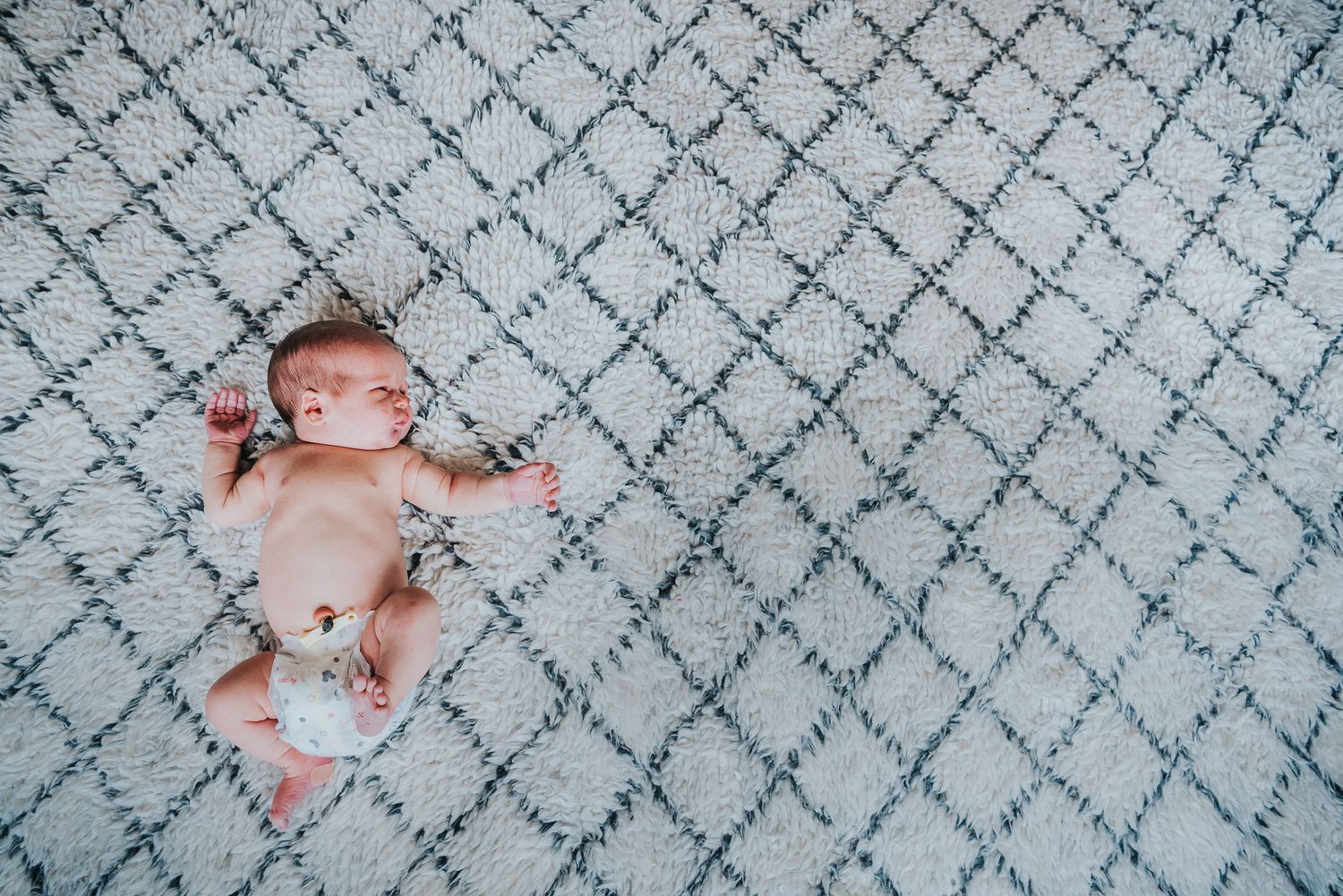 Newborn baby lays in his diaper on a white rug with navy blue diamonds.
