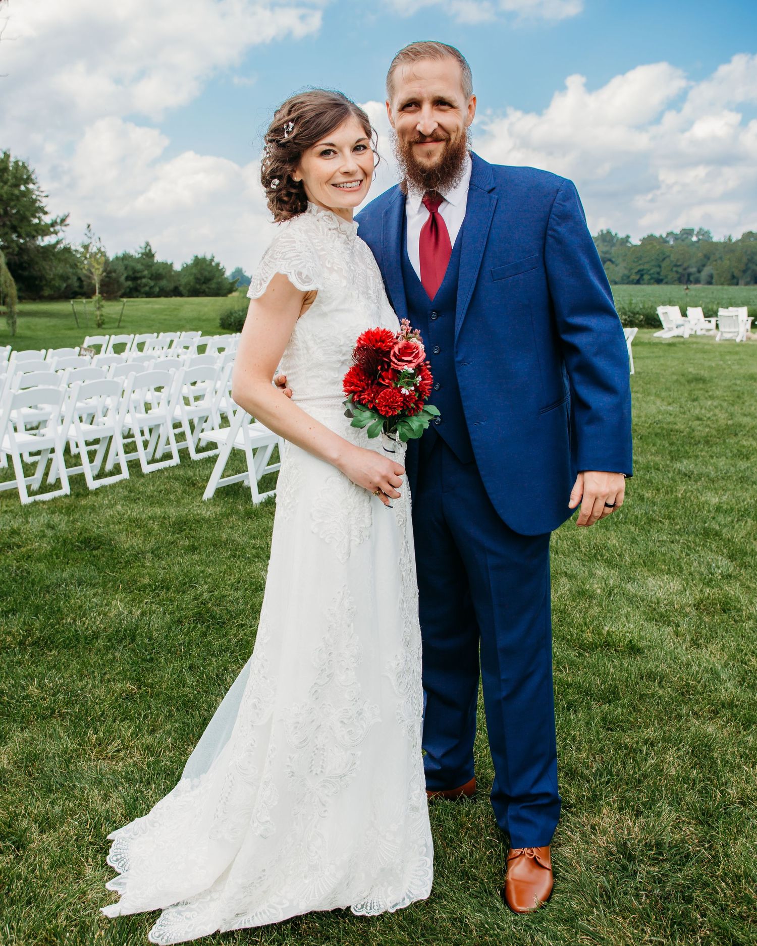 A bride in a lace gown and groom in navy suit pose on their wedding day with white chairs in background.
