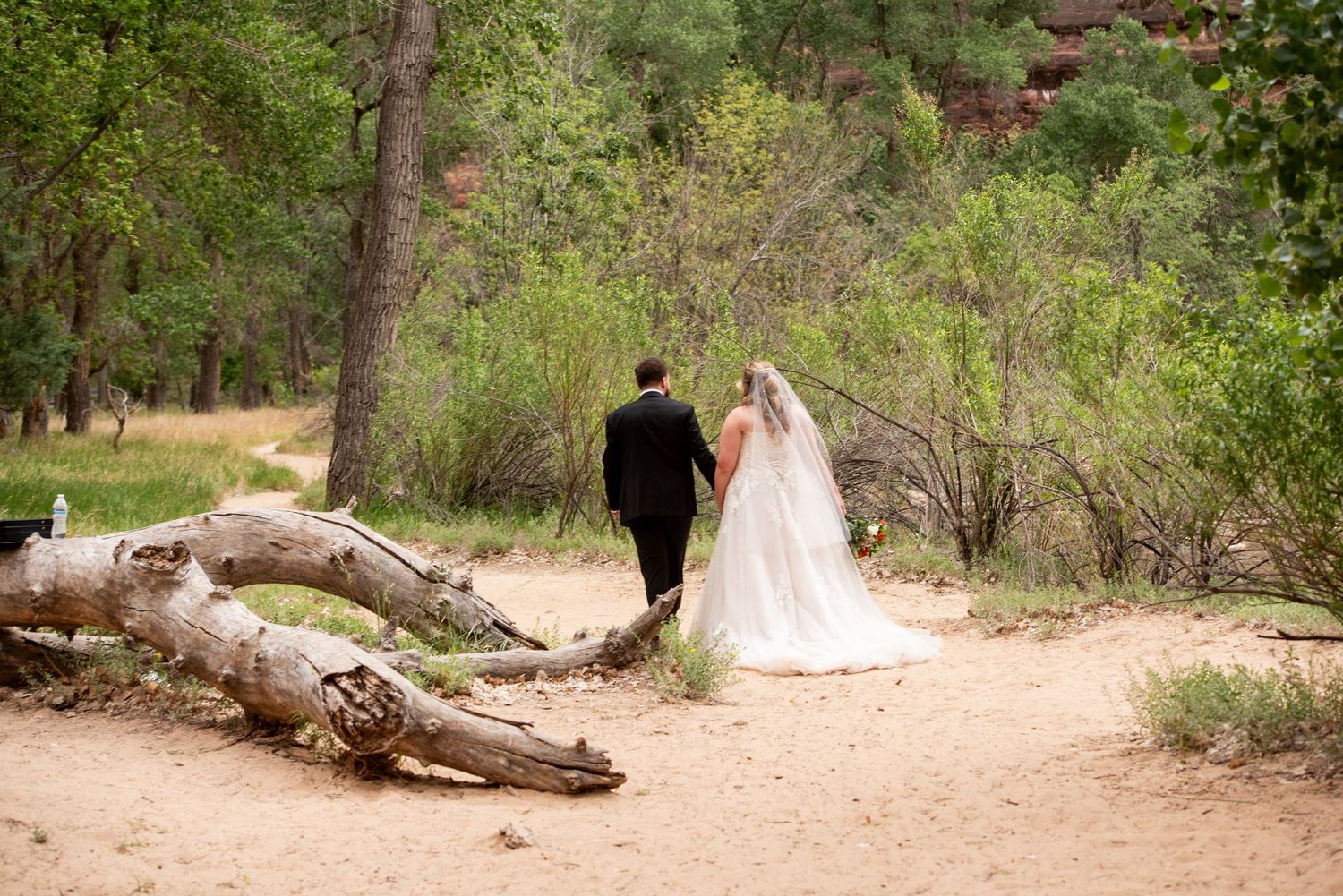 Bride and groom walking hand in hand on a sandy path through lush green forest in a scenic outdoor wedding setting.