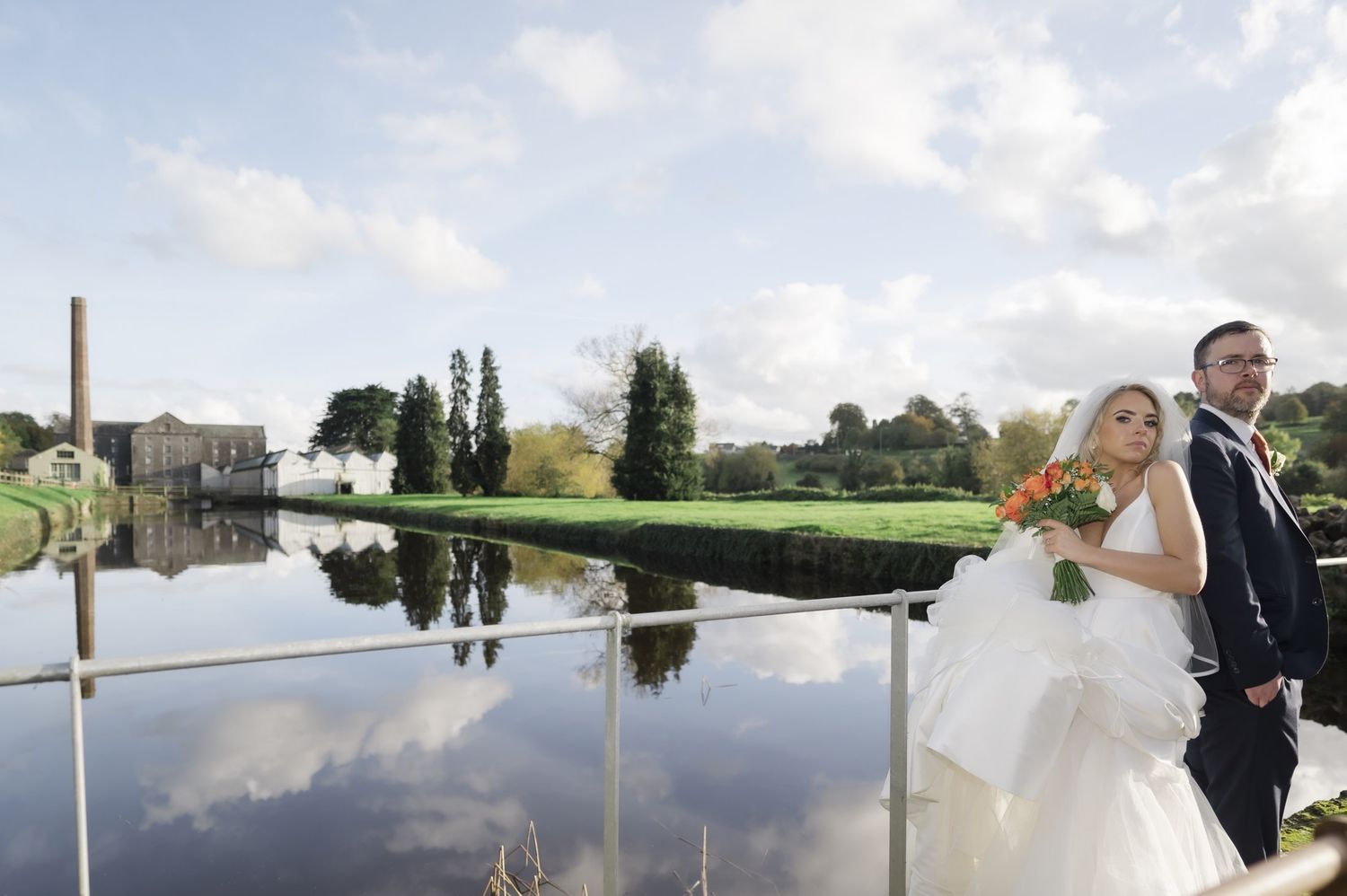 Charlie and Steven beside the water at The Millhouse, Slane, with the mill and clouds reflected behind them.