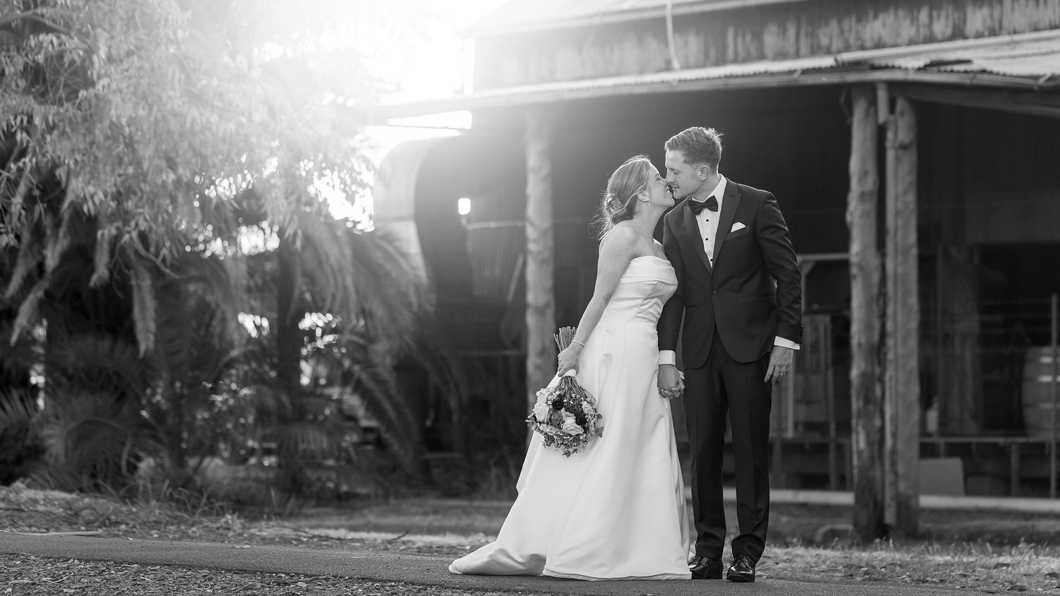 Black and white photo of a bride and groom sharing a kiss outdoors in front of a rustic wooden building at their wedding.