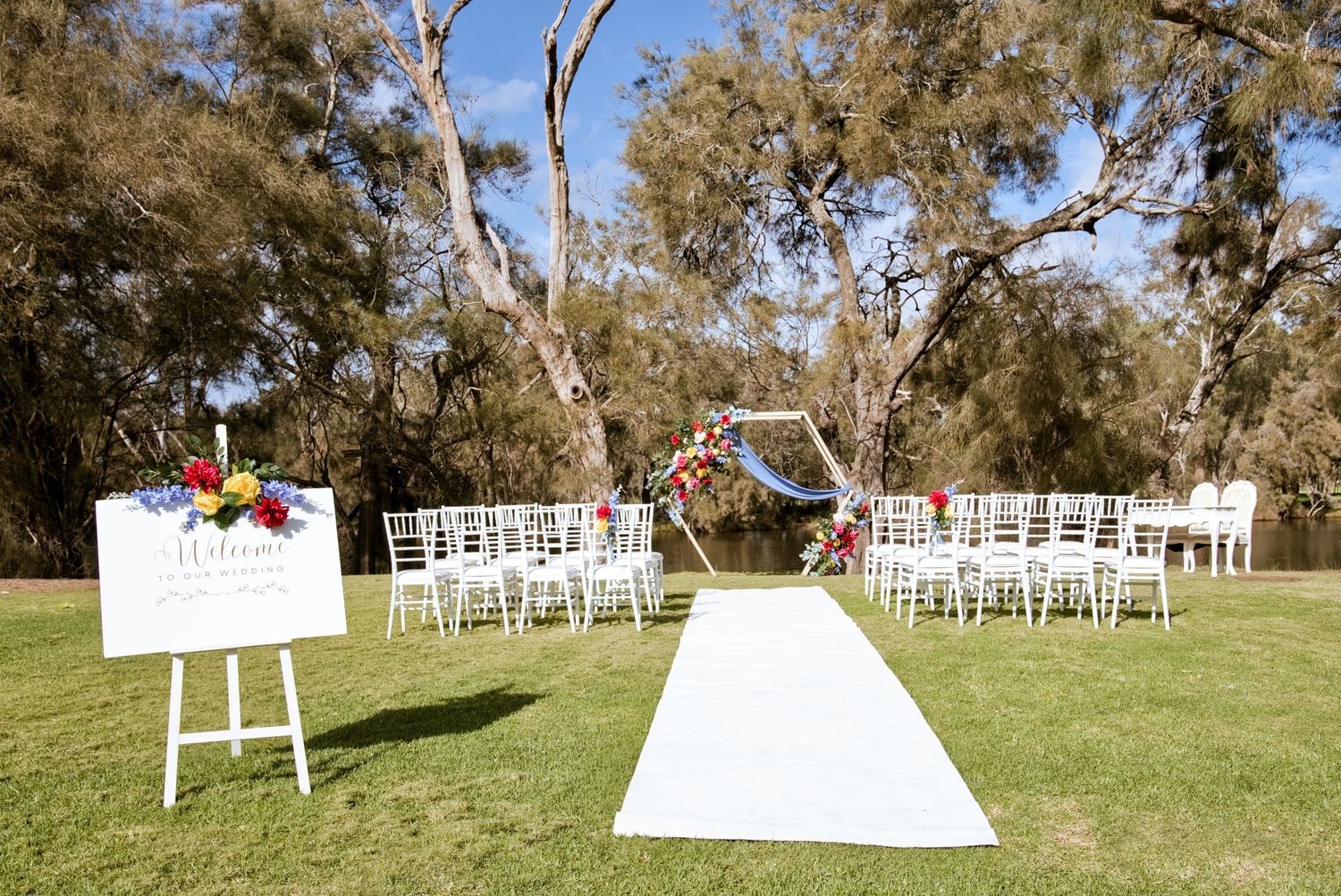 Outdoor garden wedding ceremony setup with white chairs, a floral welcome sign, white aisle runner and a floral arch beneath tall gum trees.