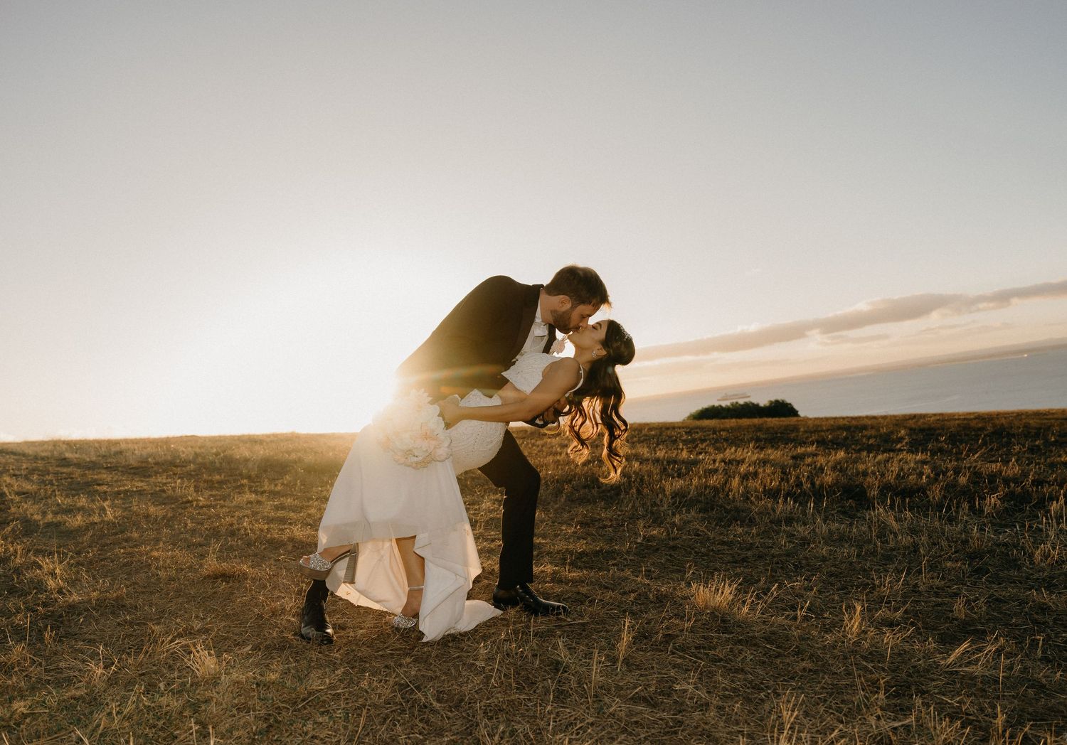 Couple sharing a romantic dip kiss at sunset in a golden field, bride in white gown and groom in black suit.
