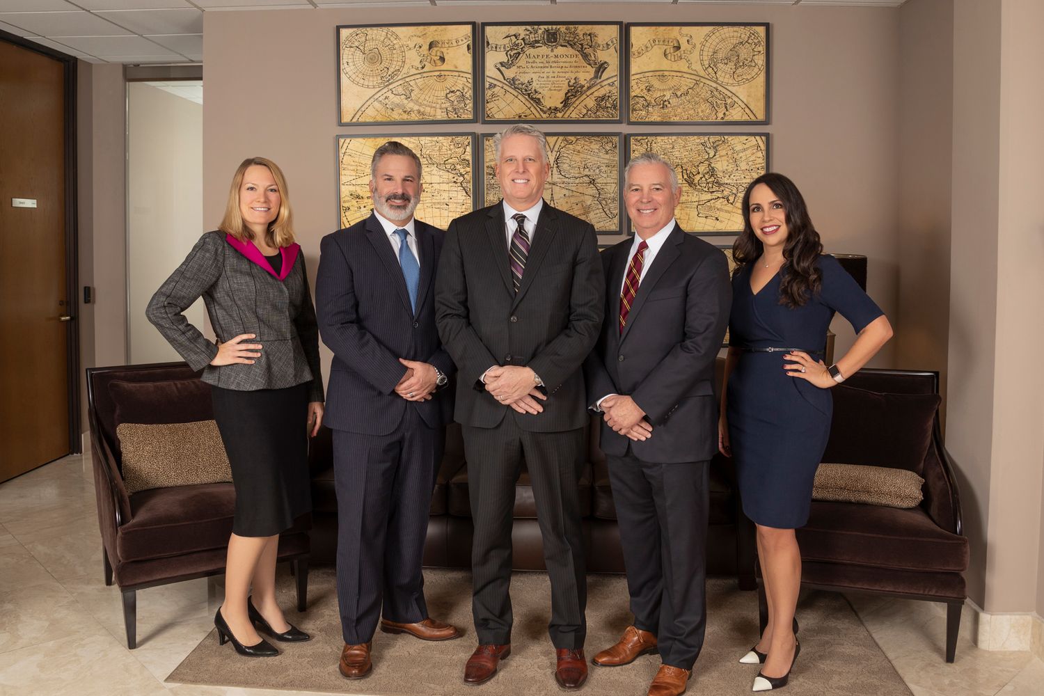 Five professionals, three men and two women in business attire, pose together in a modern office with framed maps on the wall behind them.