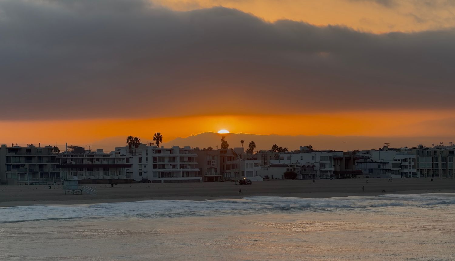 Orange sunset glowing beneath dark clouds over a quiet beachfront neighborhood with palm trees, sandy shore, and gentle ocean waves.