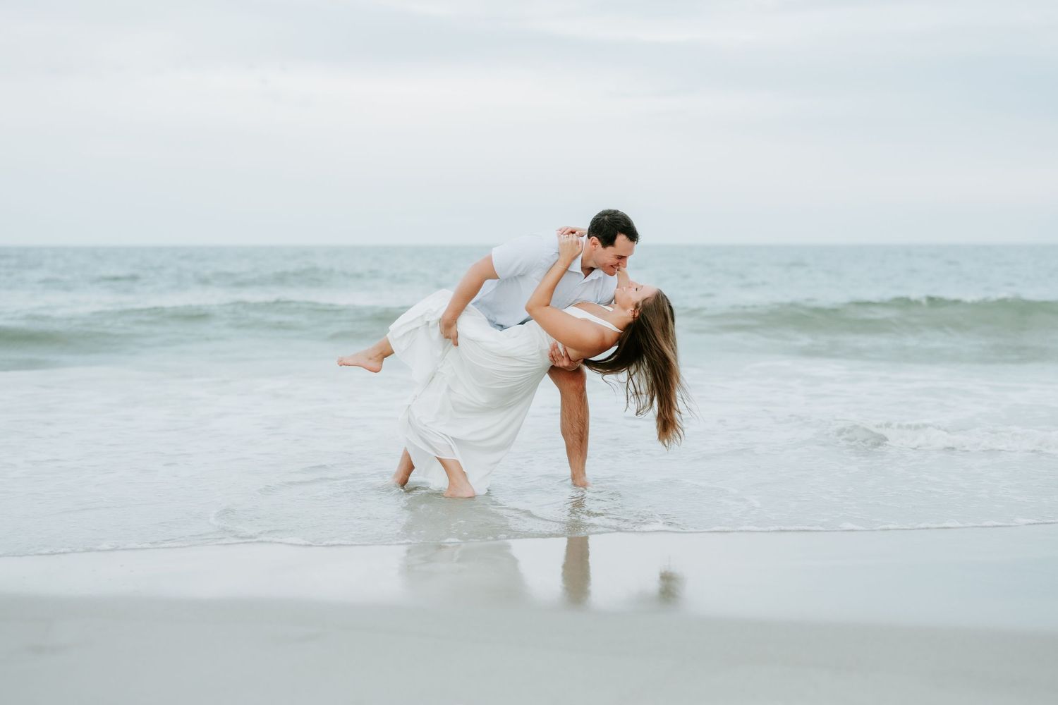 Couple sharing a romantic dip kiss on a sandy beach with ocean waves in the background.