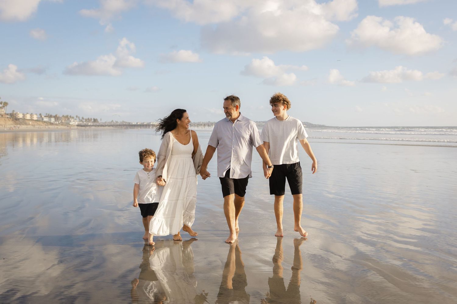 Couple walking hand in hand along a wet sandy beach at sunset while a young child in a white shirt plays nearby in the shallow water.