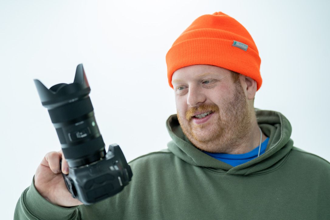 Photographer in olive hoodie and orange beanie holds and examines a professional DSLR camera with telephoto lens.