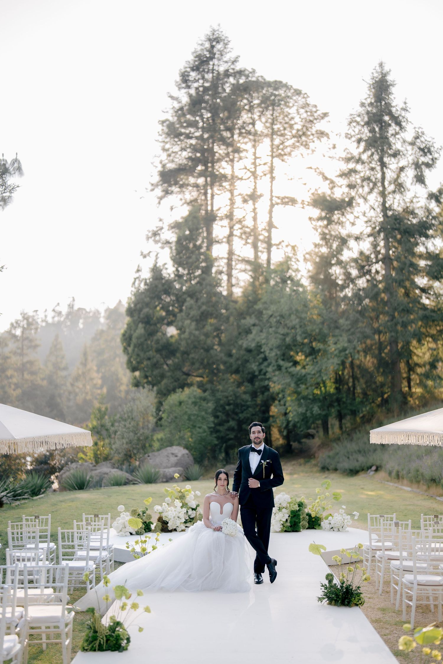 An elegant outdoor wedding ceremony setup in a wooded area with white chairs and floral arrangements at sunset.