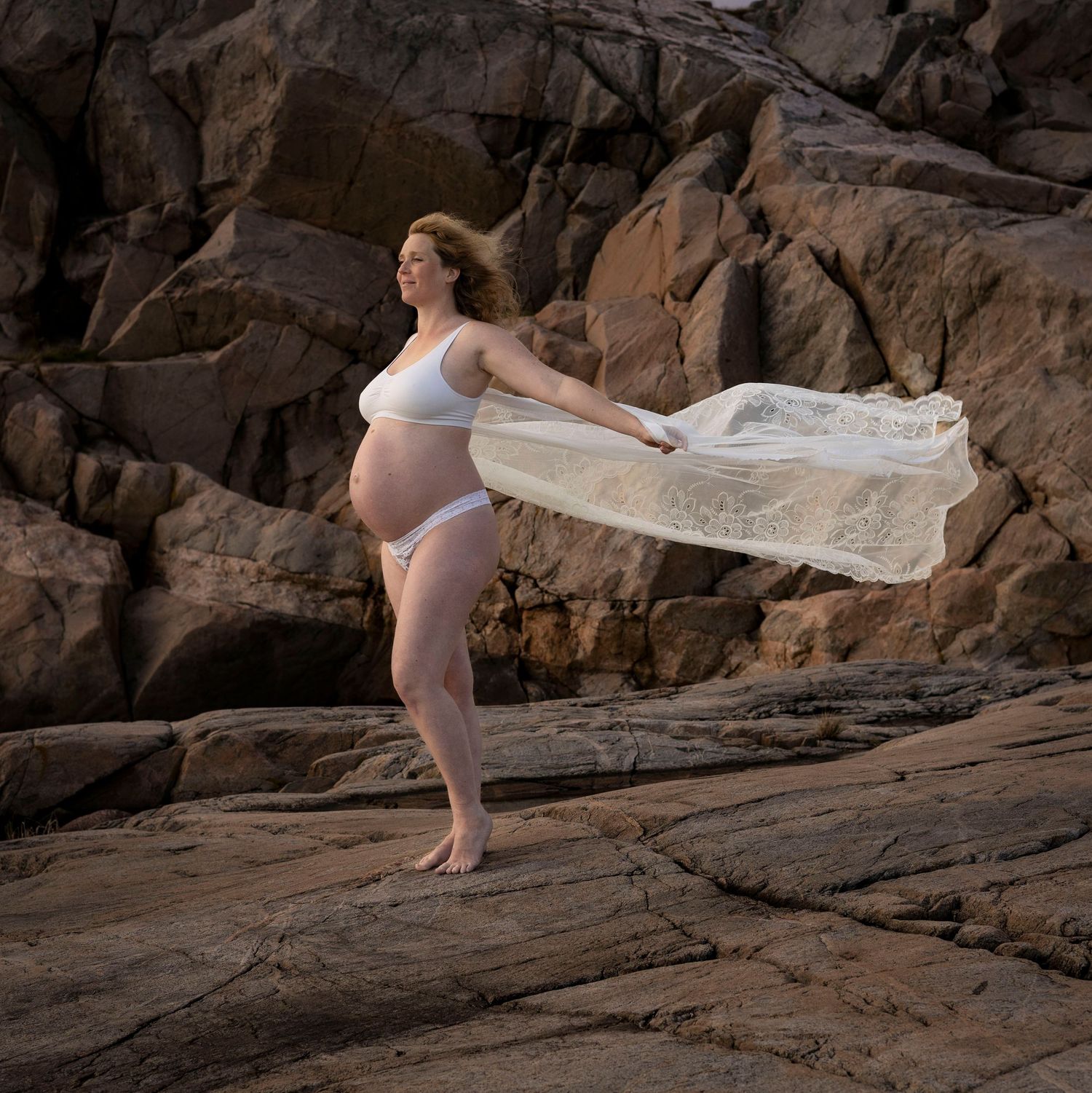 fotografering av gravid kvinna bland klipporna. hon bär en vit klänning som flyger i vinden.