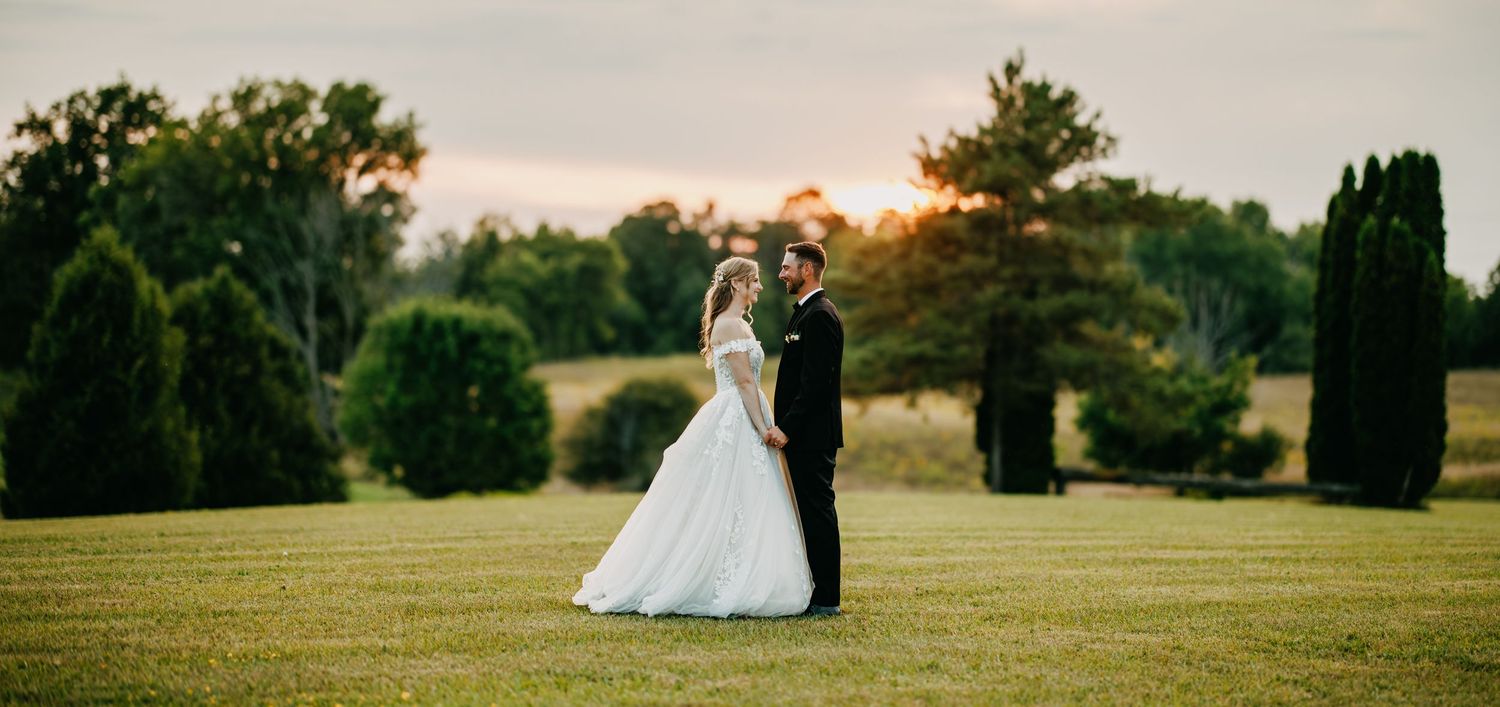 A couple in wedding attire stands together in a lush green garden setting during sunset.