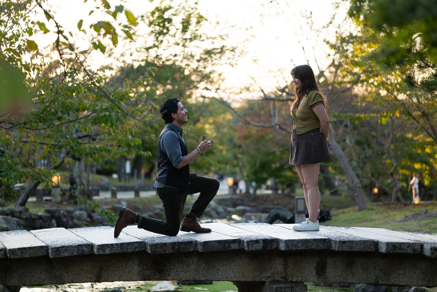 Man proposes on one knee to woman on stone bridge in autumn park.
