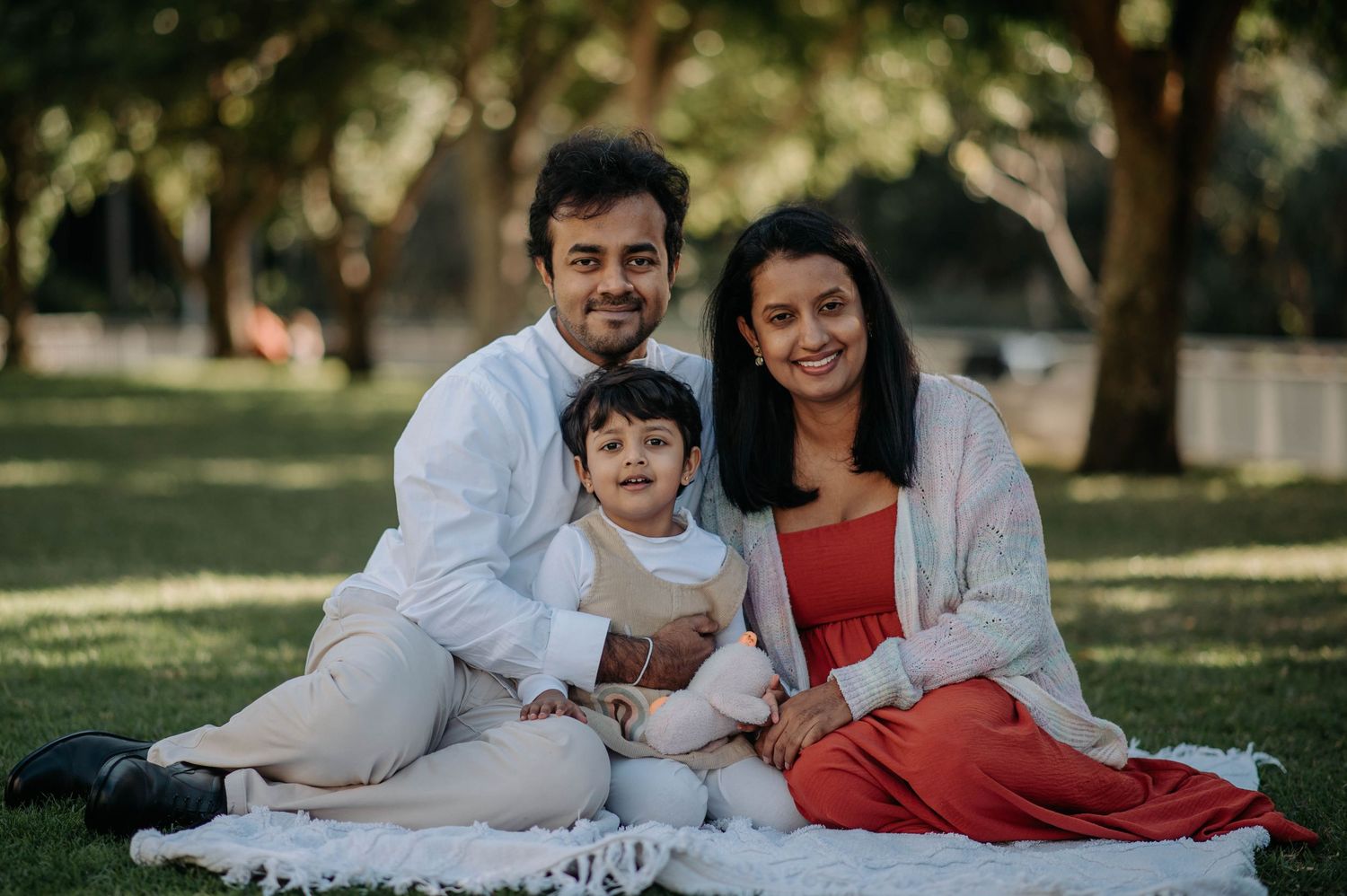 A family sits together on a white blanket outdoors in a park on a sunny day.