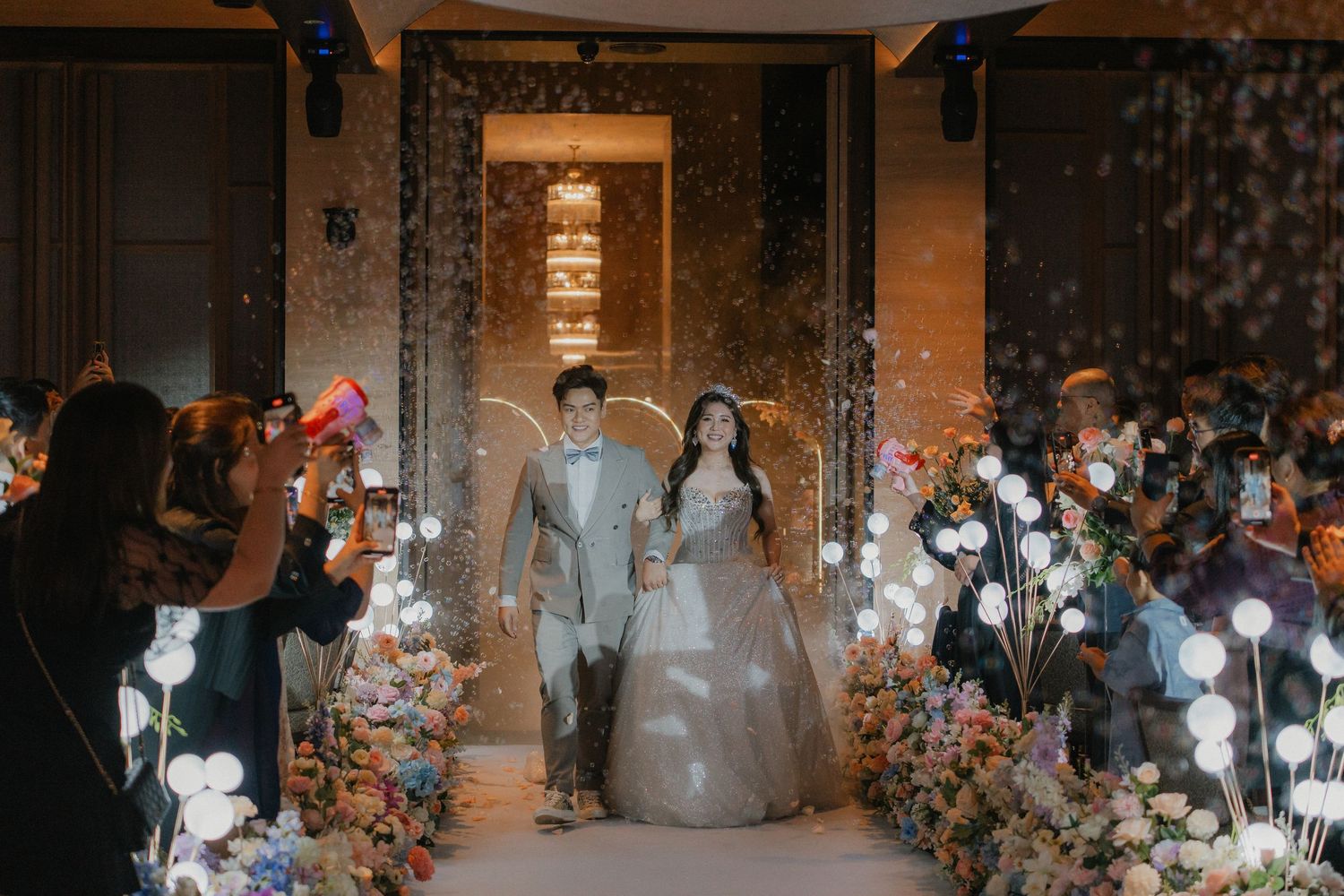 Wedding couple enters through decorative floral arch with guests lining the pathway in an elegant reception venue.
