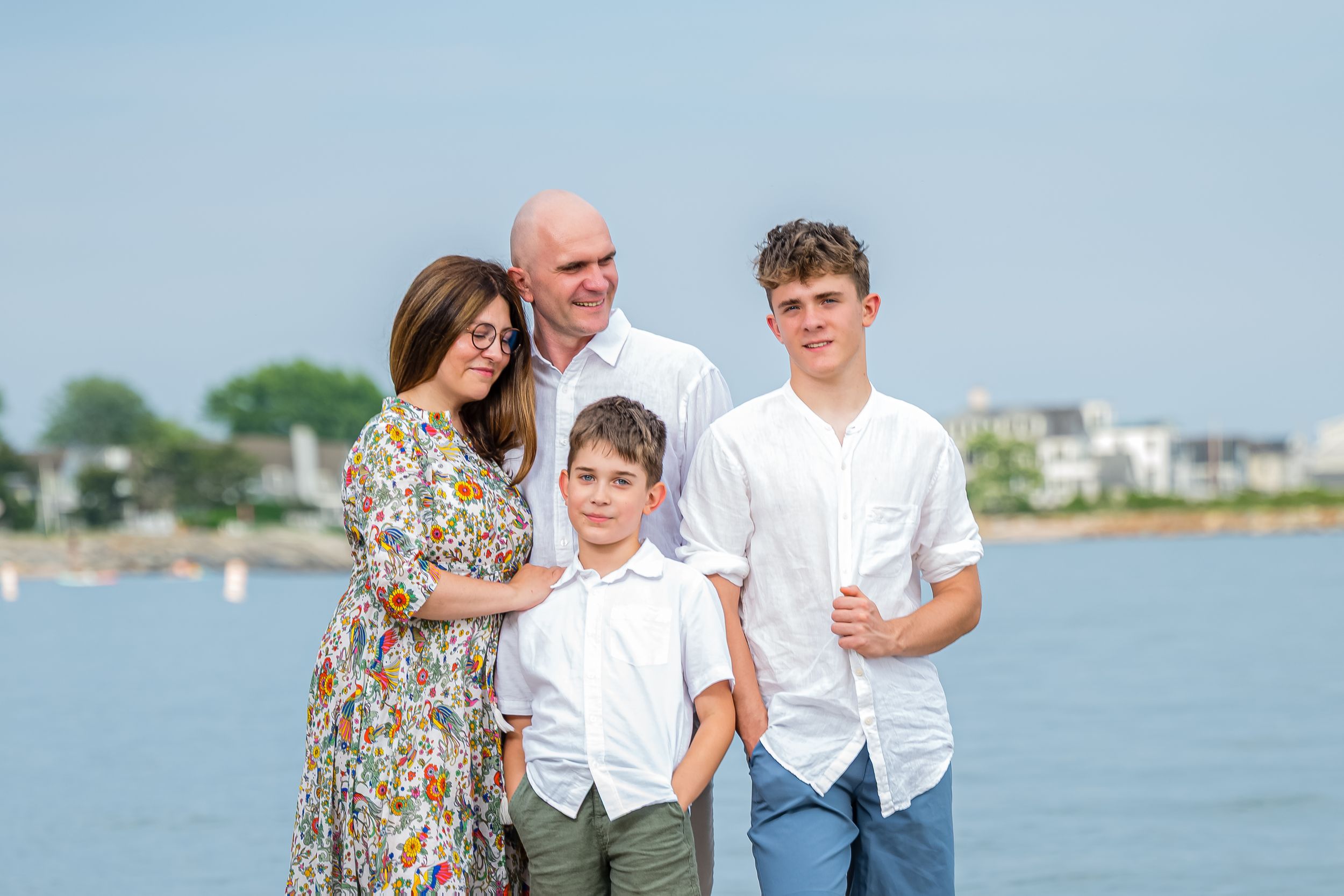 A family stands together on a beach overlooking the water with coastal homes visible in the background.