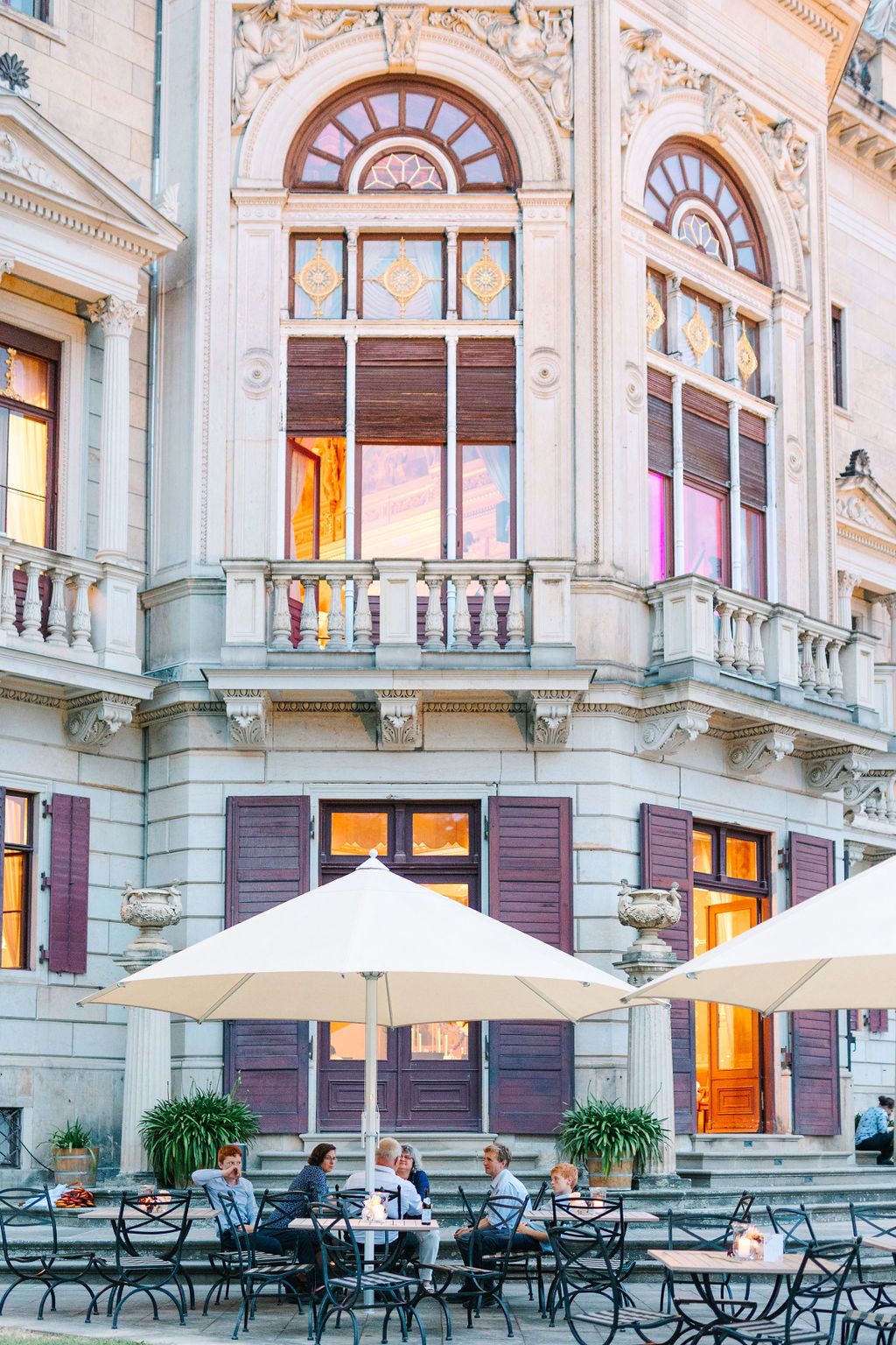 Elegant outdoor cafe terrace with white umbrellas and metal chairs against a historic building facade with ornate architecture.