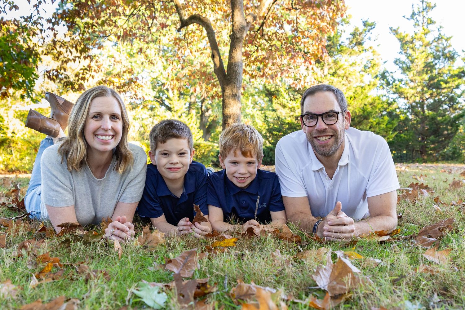Family lying on autumn ground covered in fallen leaves during outdoor photo session in park.