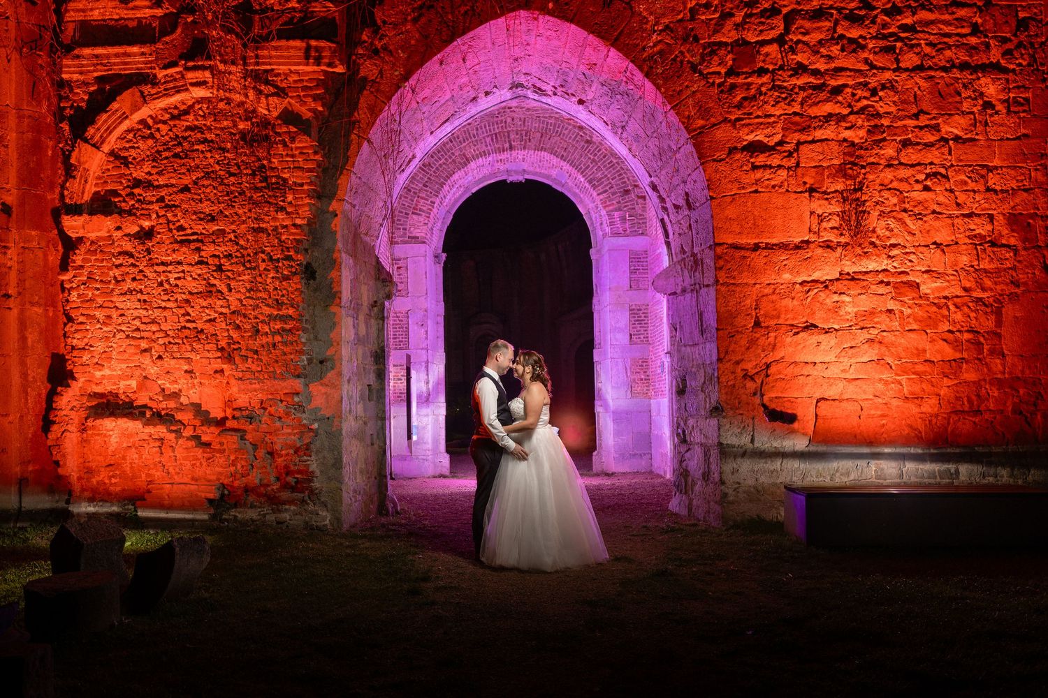 Wedding couple illuminated in vibrant pink and orange lighting within historic stone arch tunnel.