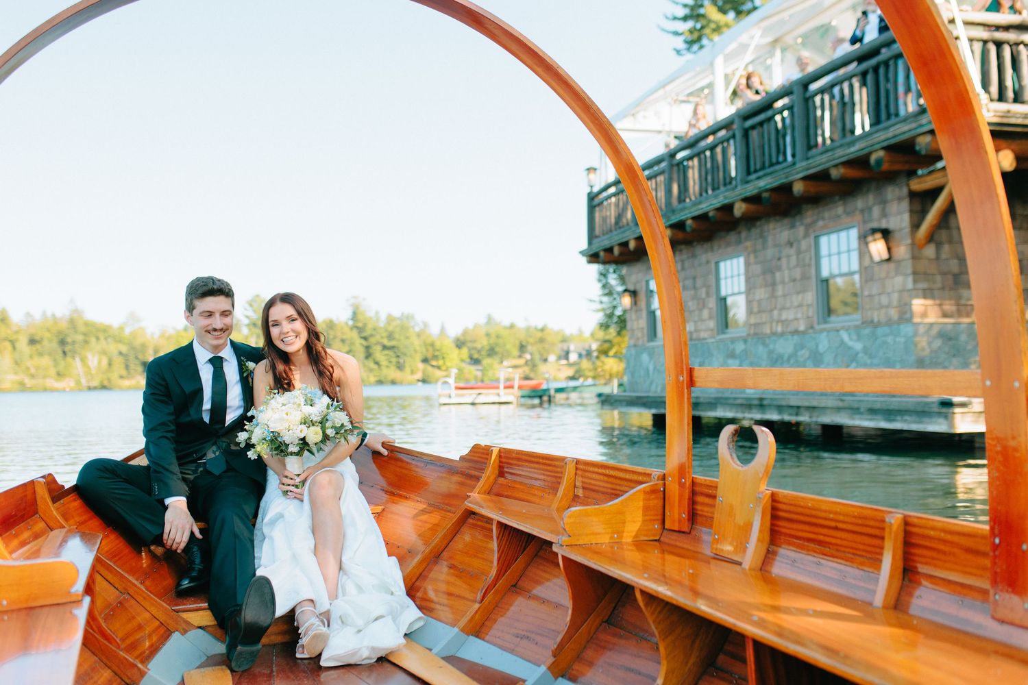 A couple sits together in a wooden boat with orange oars on a lake near a building with a balcony.