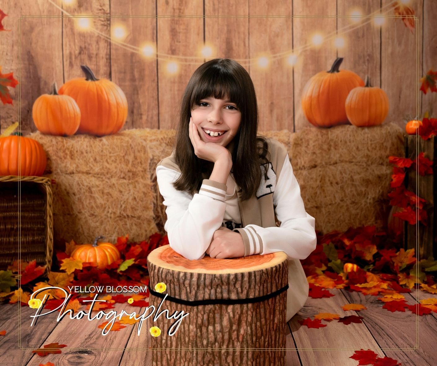 Fall themed portrait on wooden backdrop with hay bales, pumpkins, autumn leaves and string lights.