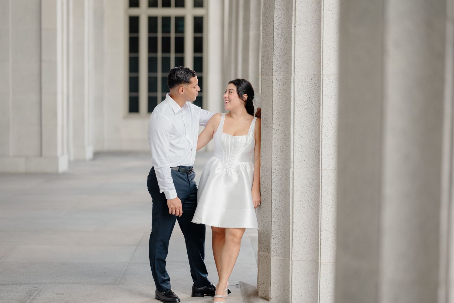 A couple in wedding attire shares an intimate moment among elegant white columns of a grand building.