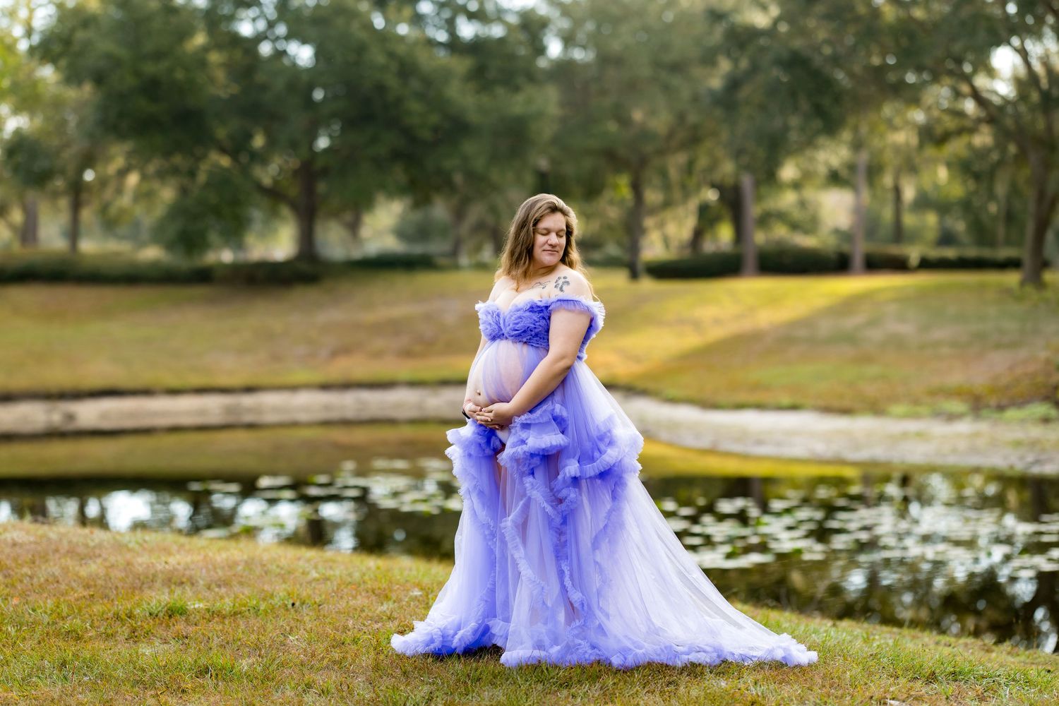 A person in a flowing lavender gown poses gracefully near a serene pond surrounded by autumn trees.