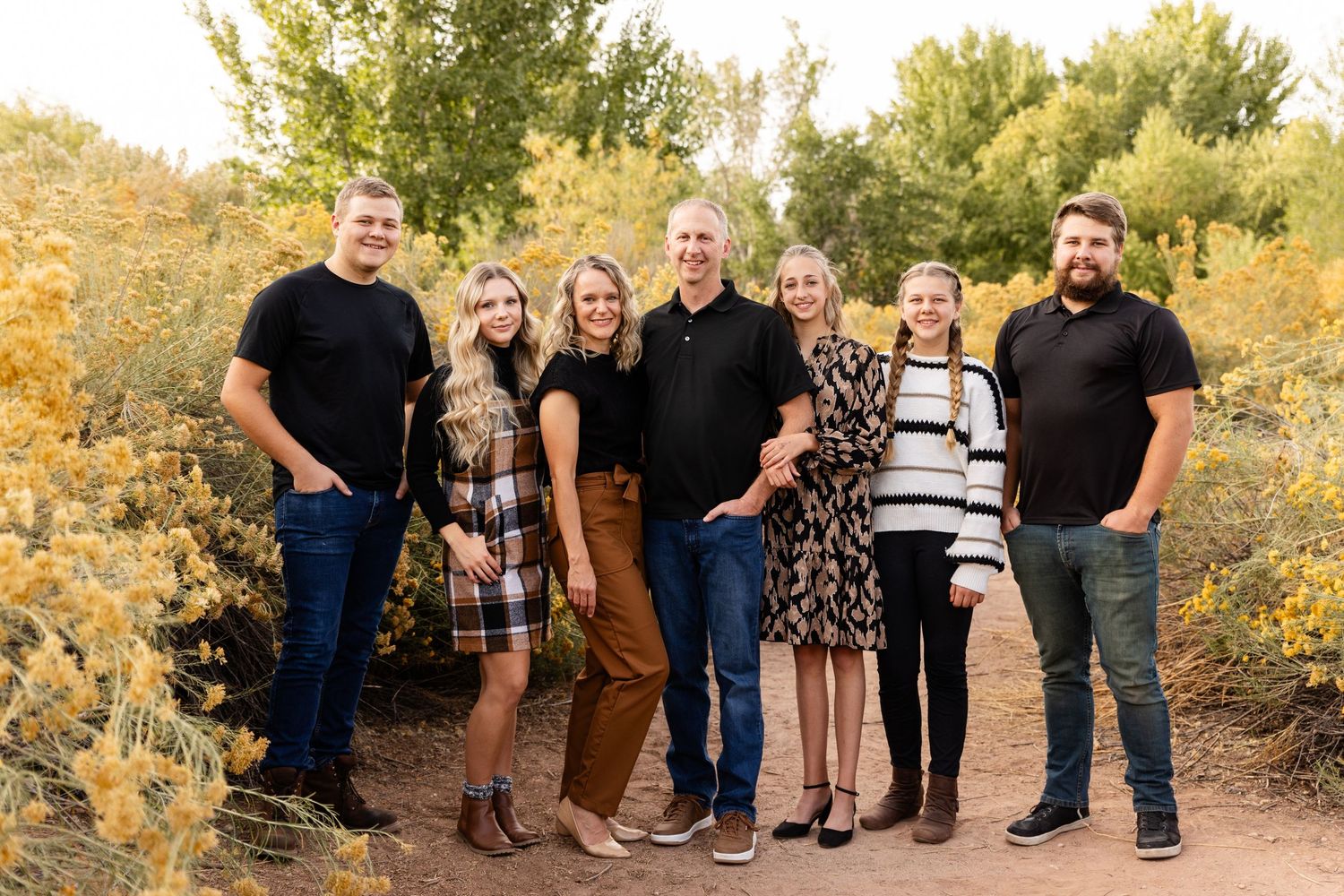 Family of 7. 3 girls and 2 boys (all adults) smiling at the camera wearing black, orange white and plaid fall colors with faded yellow bloom