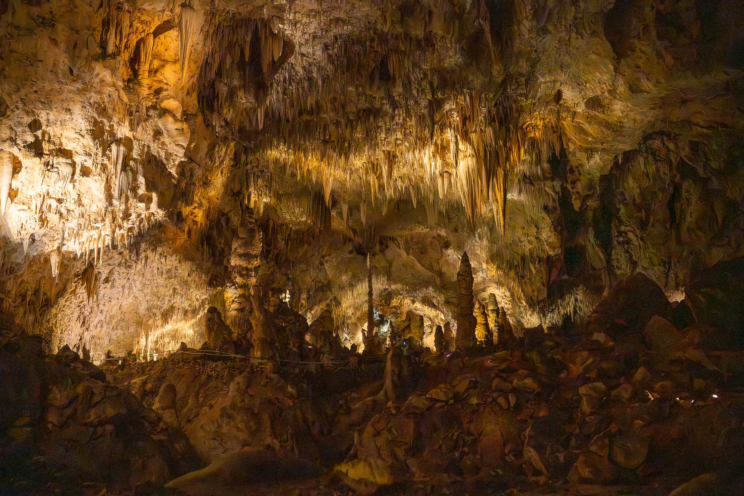 Dramatic limestone formations and stalactites illuminated in golden light inside a natural cave chamber.
