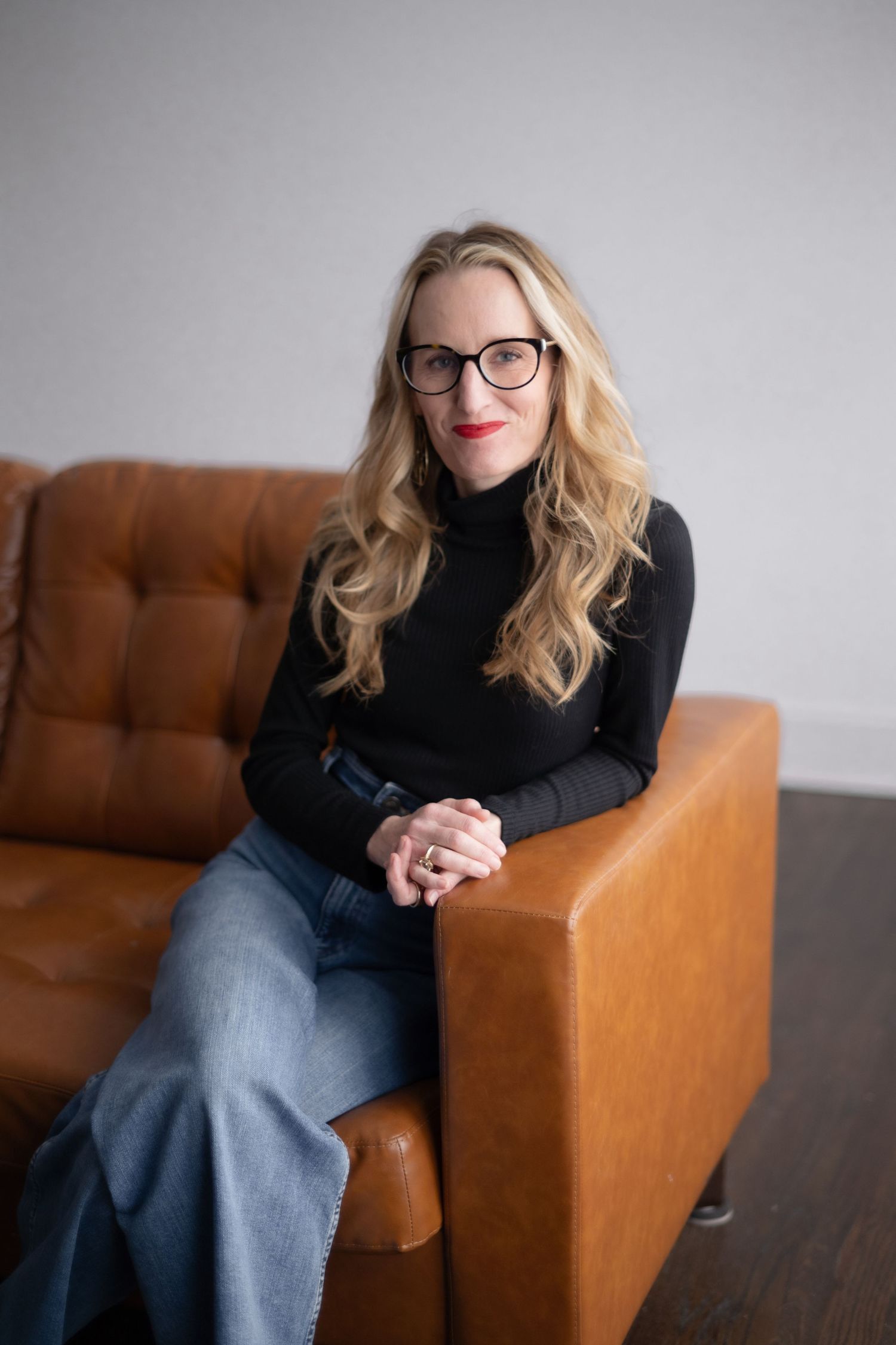 Professional portrait series showing person in black sweater and glasses seated on brown leather chair in studio setting.