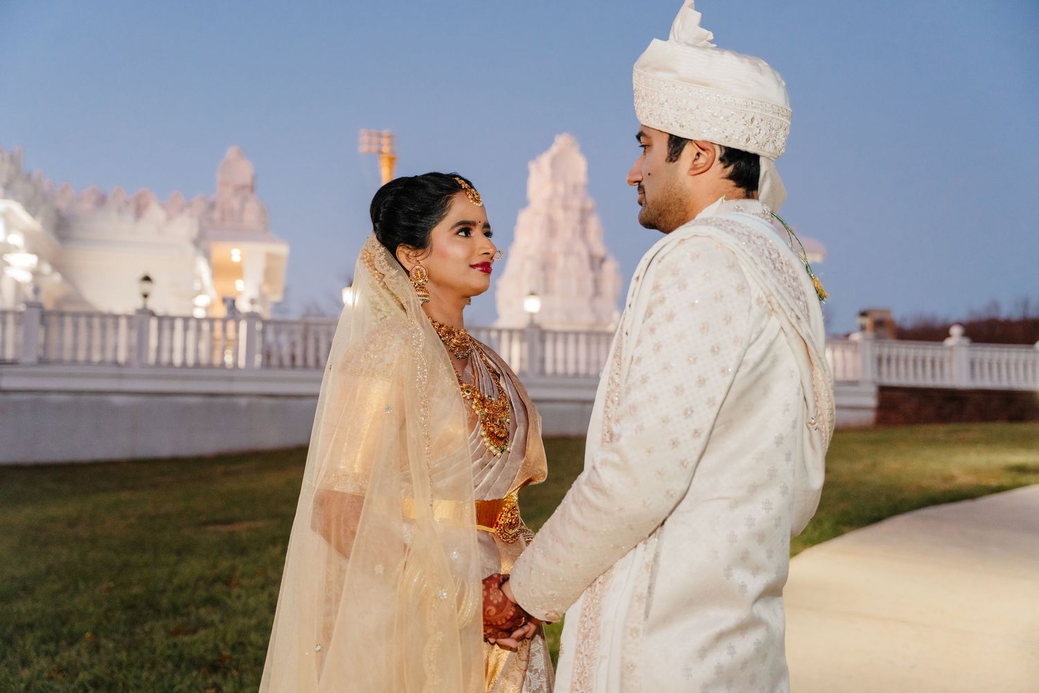 A couple in traditional Indian wedding attire stands together outdoors during their celebration at sunset.