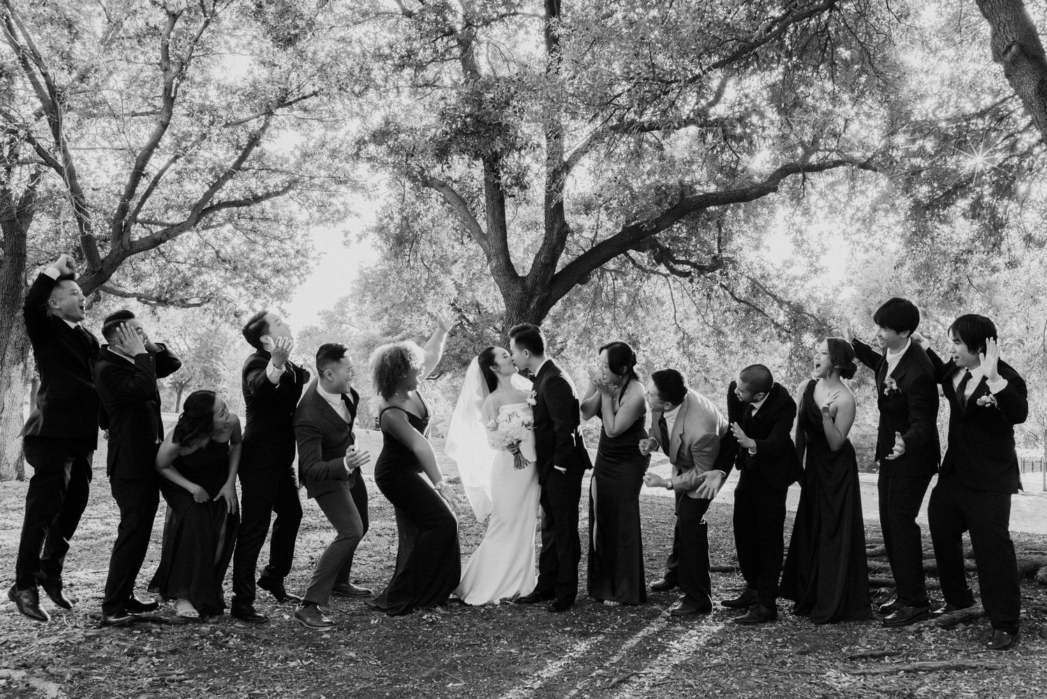 Black and white wedding party photo with bride and groom kissing under trees.