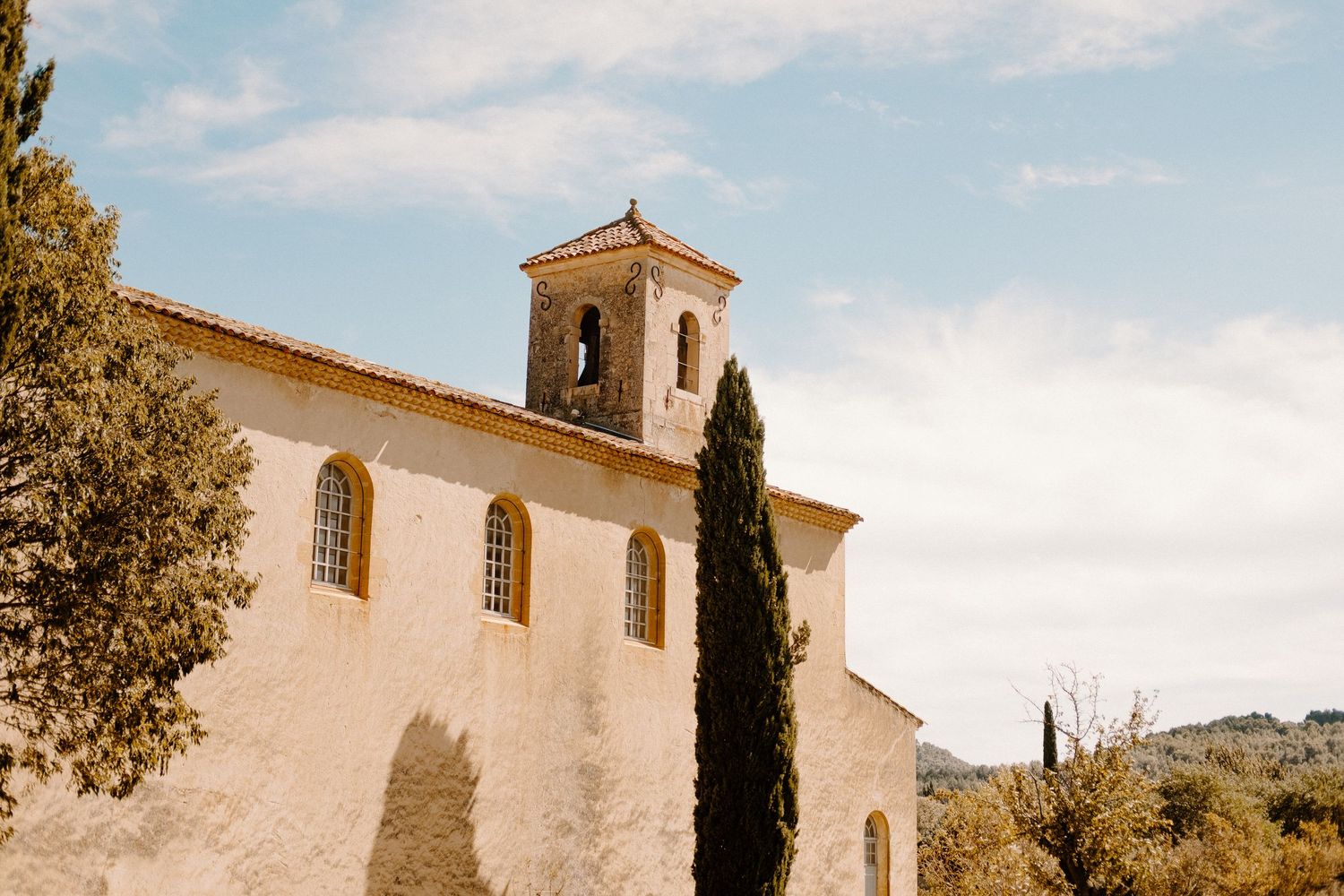 A white Mediterranean church with cypress trees and a bell tower stands against a blue sky in a rustic setting.