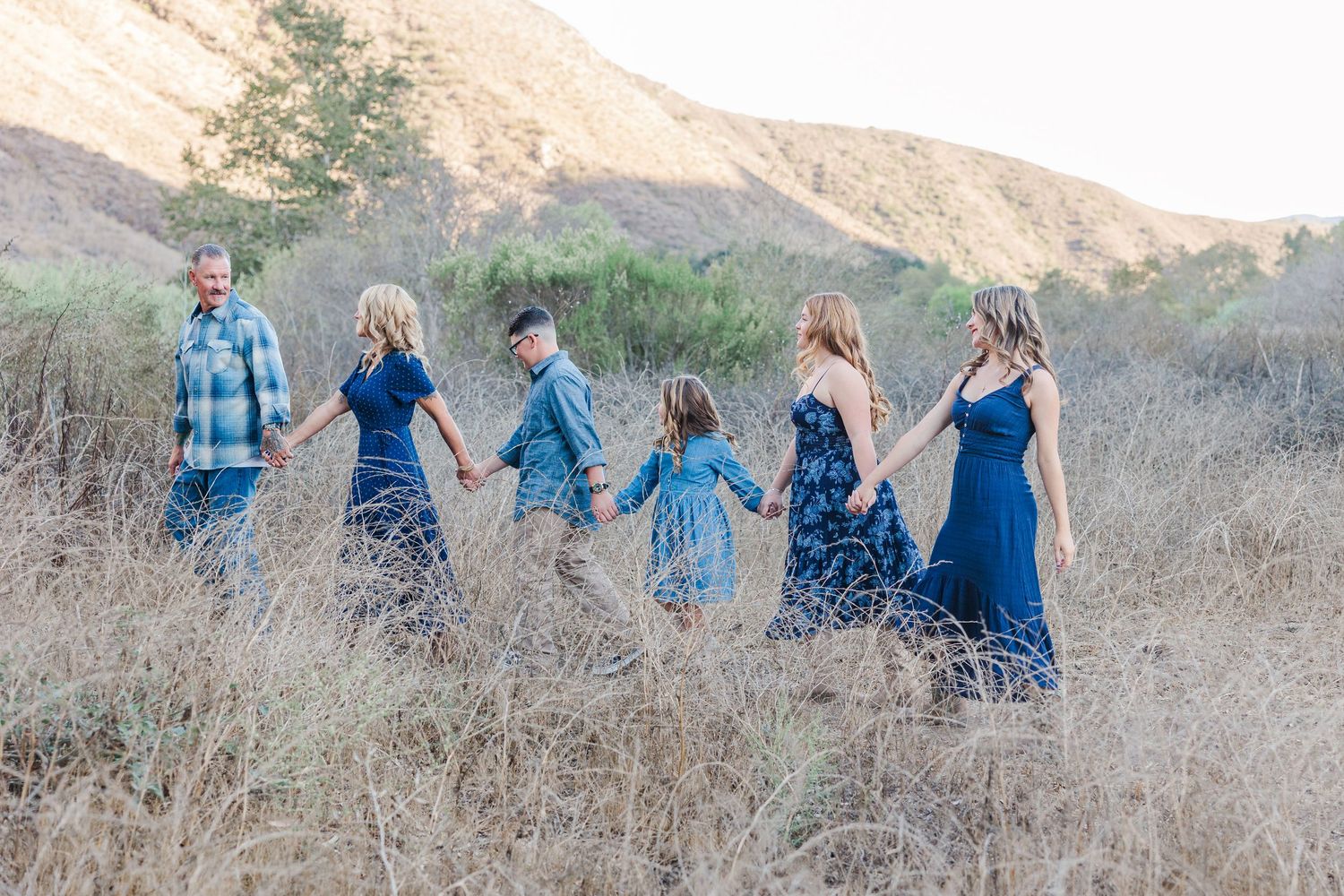 A group in coordinating blue outfits holds hands while walking through a golden hillside at sunset.