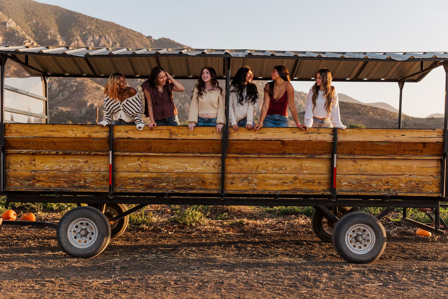 A wooden hay wagon with large wheels sits parked in a rustic farm setting at sunset.