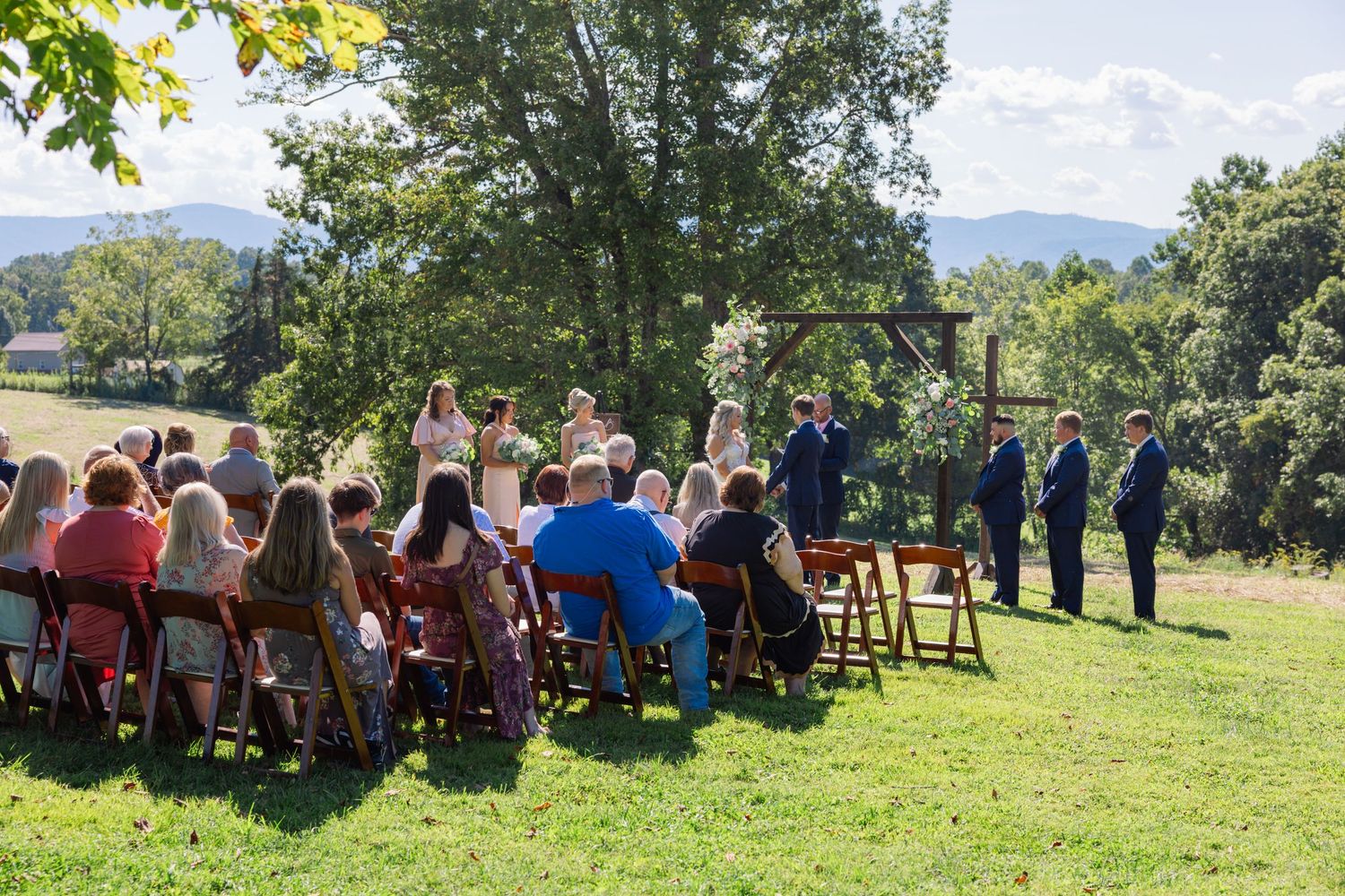 Panoramic view of outdoor wedding ceremony guests seated in wooden chairs on a grassy hillside under blue summer skies.