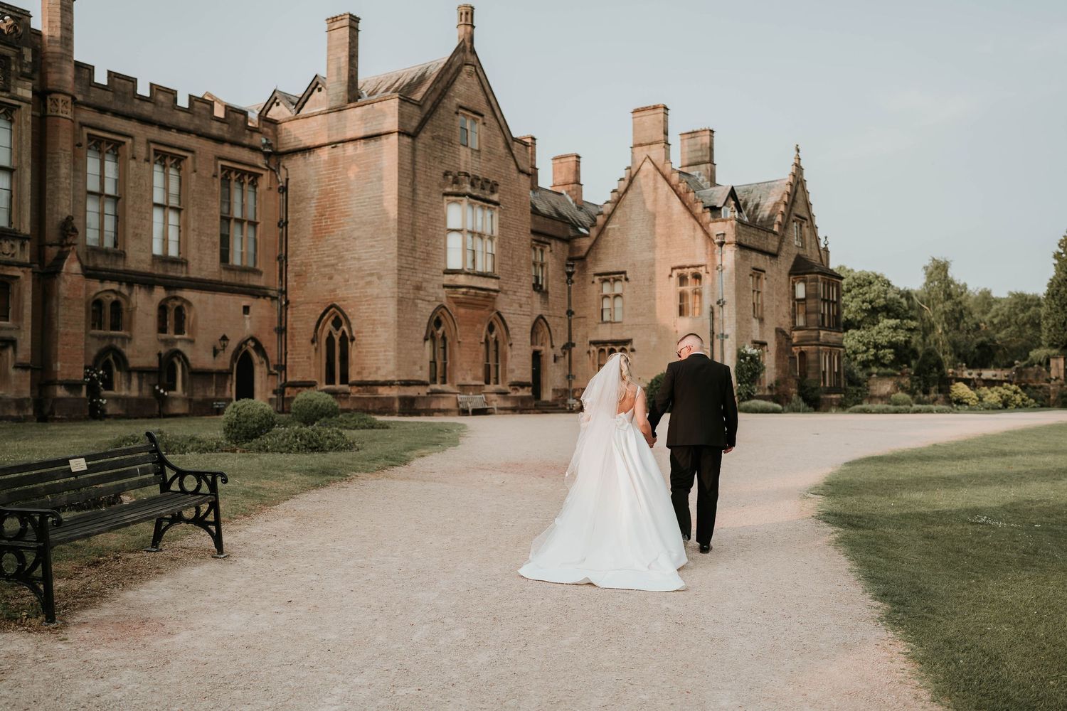 A wedding couple walks along a stone path outside a Newstead abbey with ornate architecture and chimneys.
