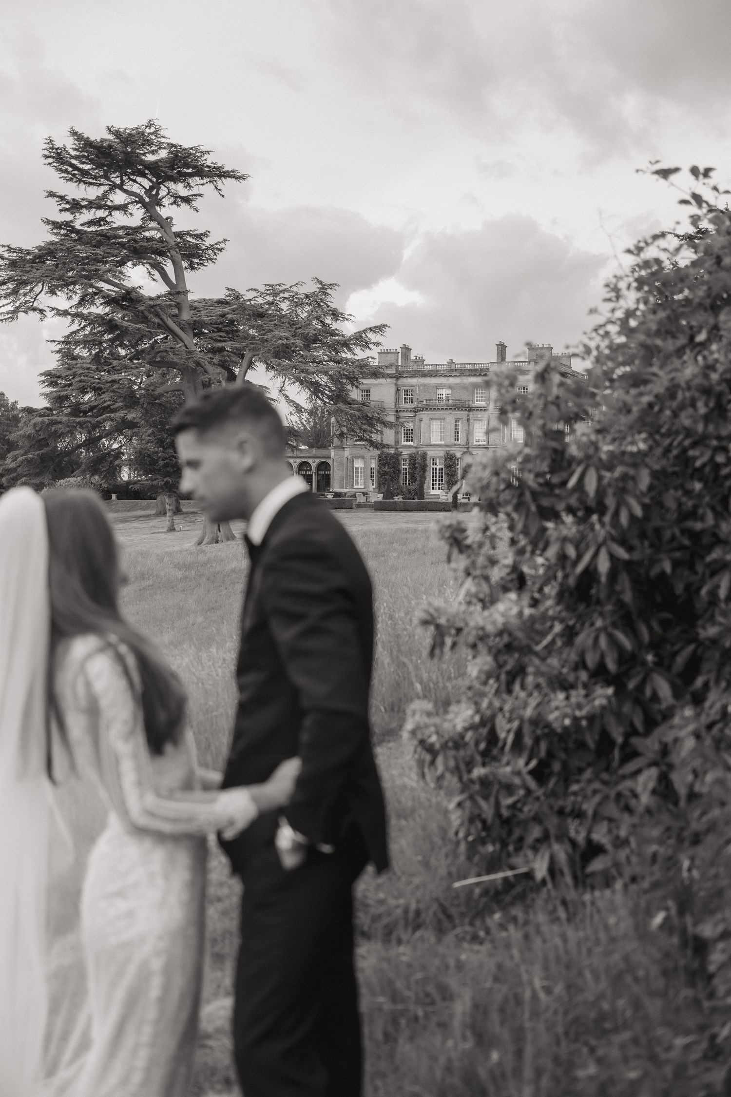 A black and white wedding photo shows a couple in formal attire standing near a grand estate with lush gardens.