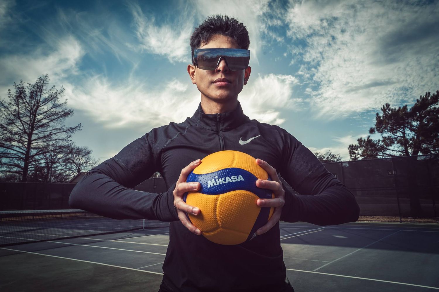 A person holds a volleyball outdoors on a court against a dramatic cloudy sky at sunset.