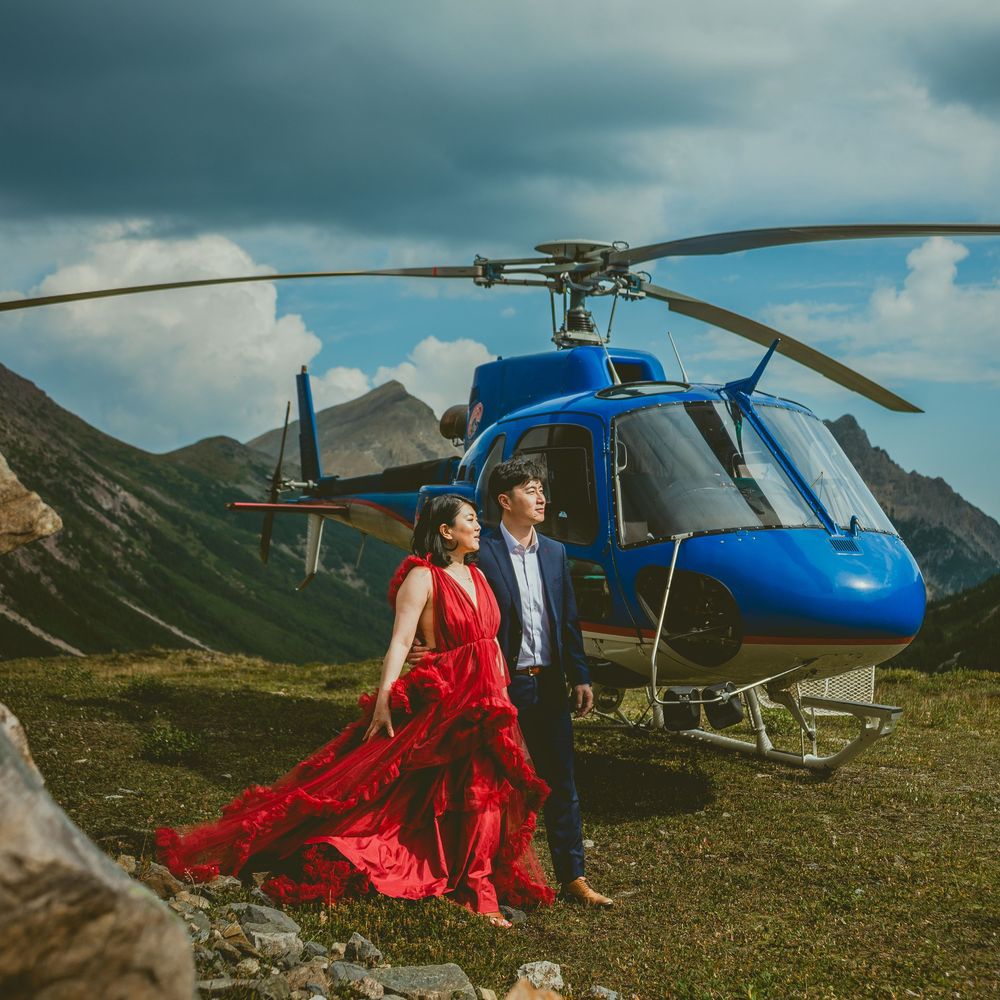 Helicopter parked in dramatic mountain landscape with moody storm clouds and bride in flowing red dress.